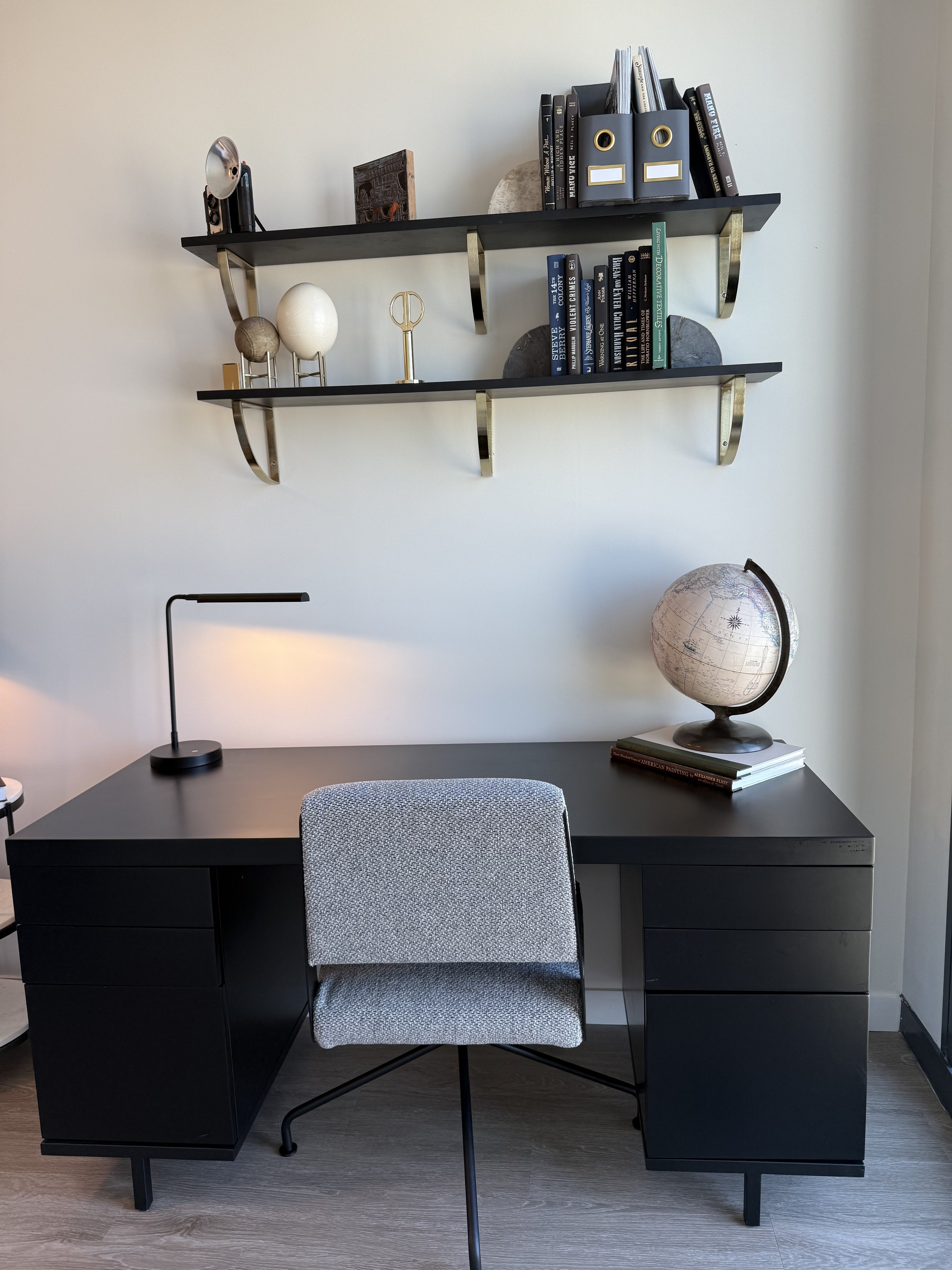 Modern black desk with grey chair, black table lamp, globe on books, and wall-mounted shelves with decorative items and books.