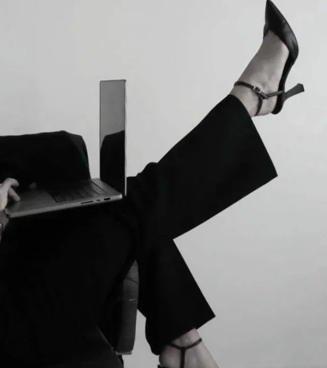 Person sitting with legs crossed on a chair, using a laptop and a mirror held in their lap, with the laptop and mirror creating an illusion of a person's legs and feet wearing high heels.