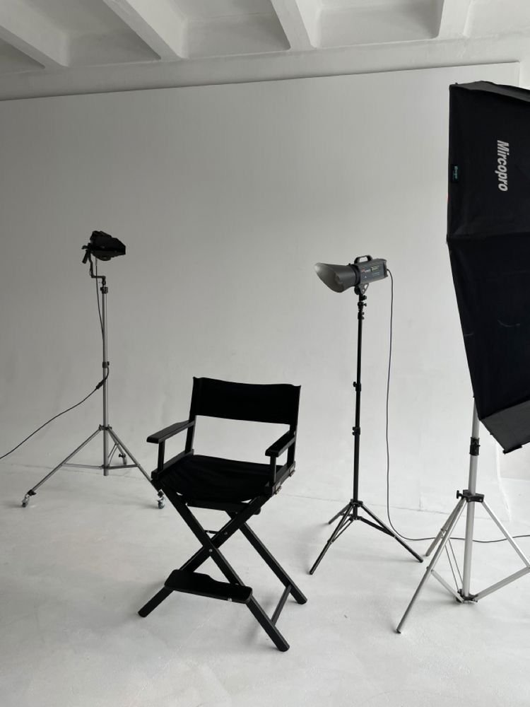 Photography studio setup with director's chair, softbox light, spotlights, and backdrop on a white floor and wall.