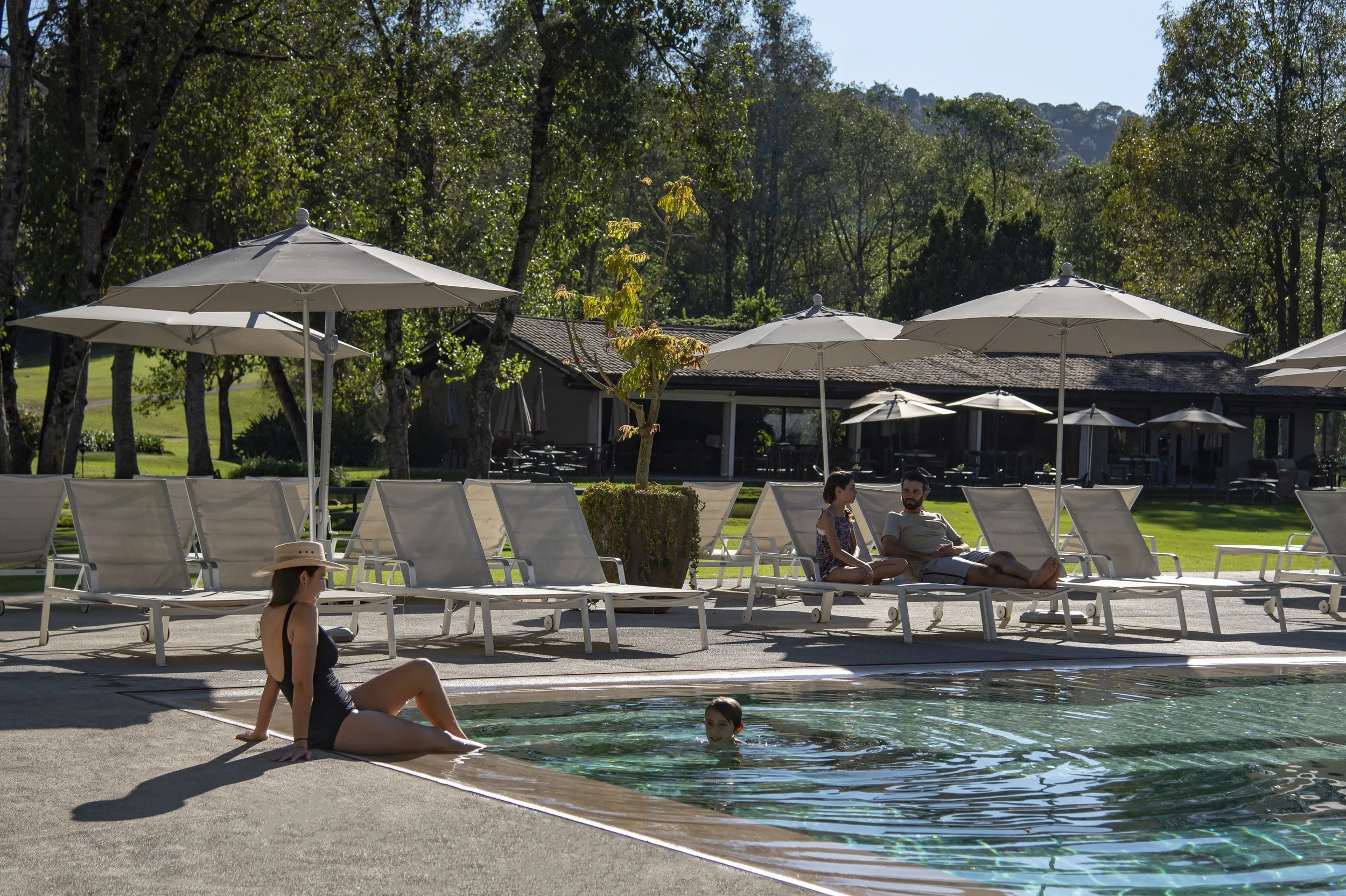 People relaxing by a swimming pool with lounge chairs and umbrellas, surrounded by trees and a building in the background.