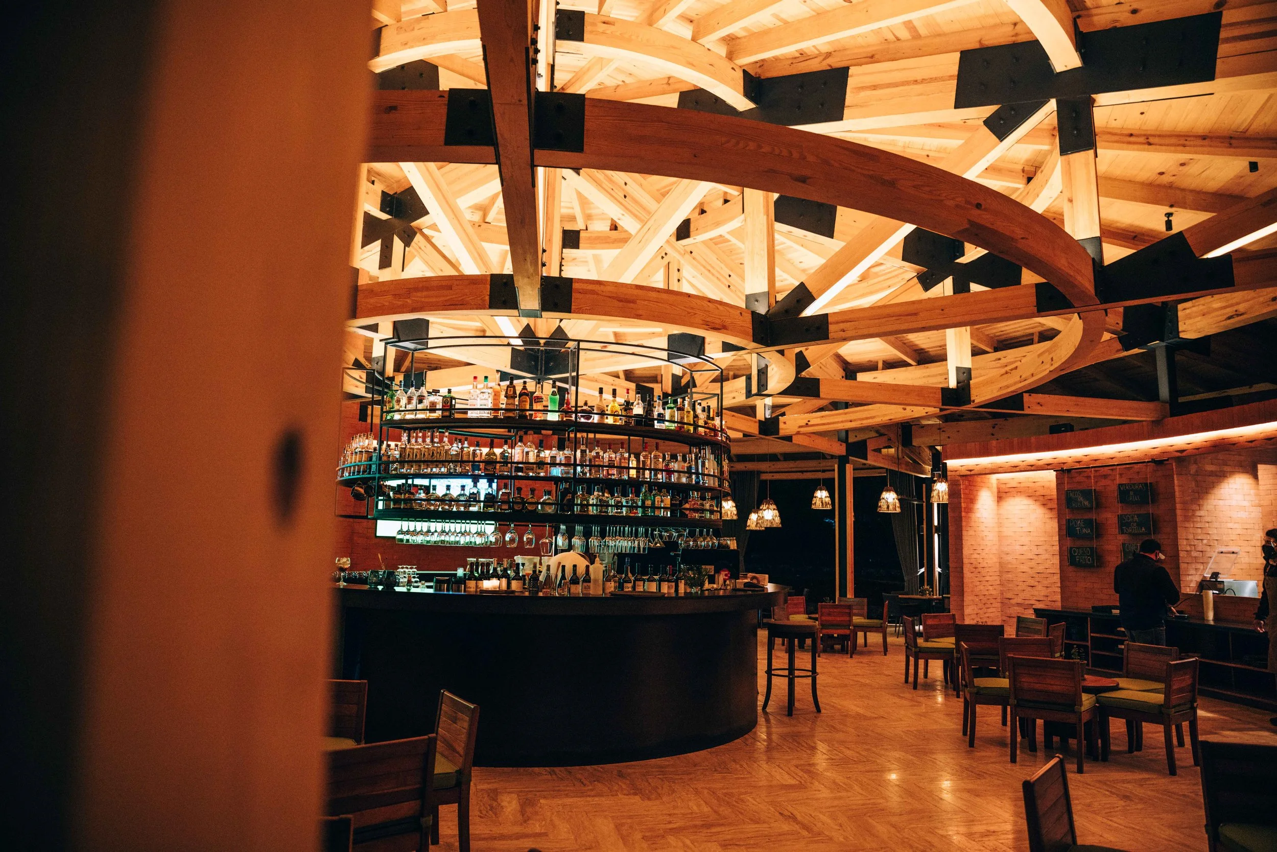 Interior of a modern bar or restaurant with a wooden ceiling featuring intricate curved beams, a circular bar with bottles of liquor and hanging glasses, and wooden tables and chairs. There are hanging pendant lights and a brick wall with menu boards