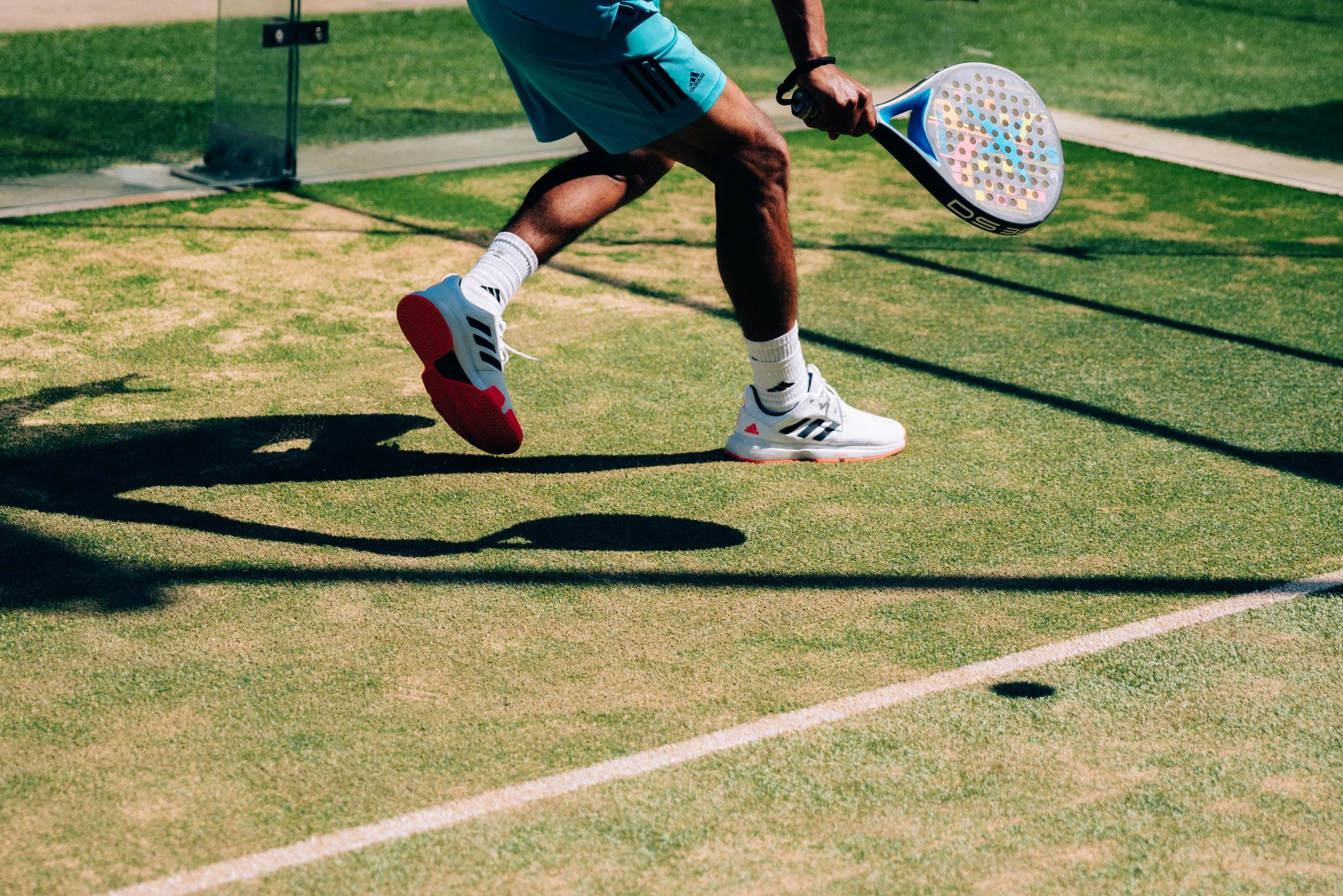 A tennis player wearing white Adidas sneakers and white socks on a grass tennis court, preparing to hit a tennis ball with a racquet.