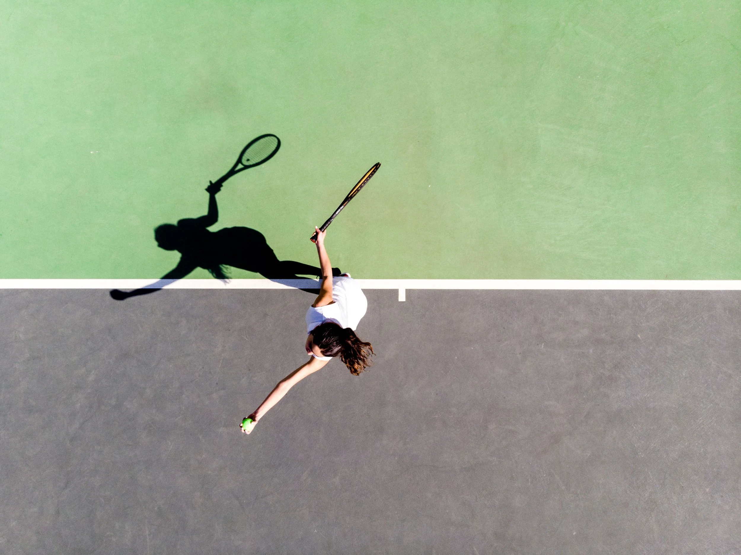 A woman playing tennis on an outdoor court, serving the ball with a tennis racket, with her shadow cast on the court.