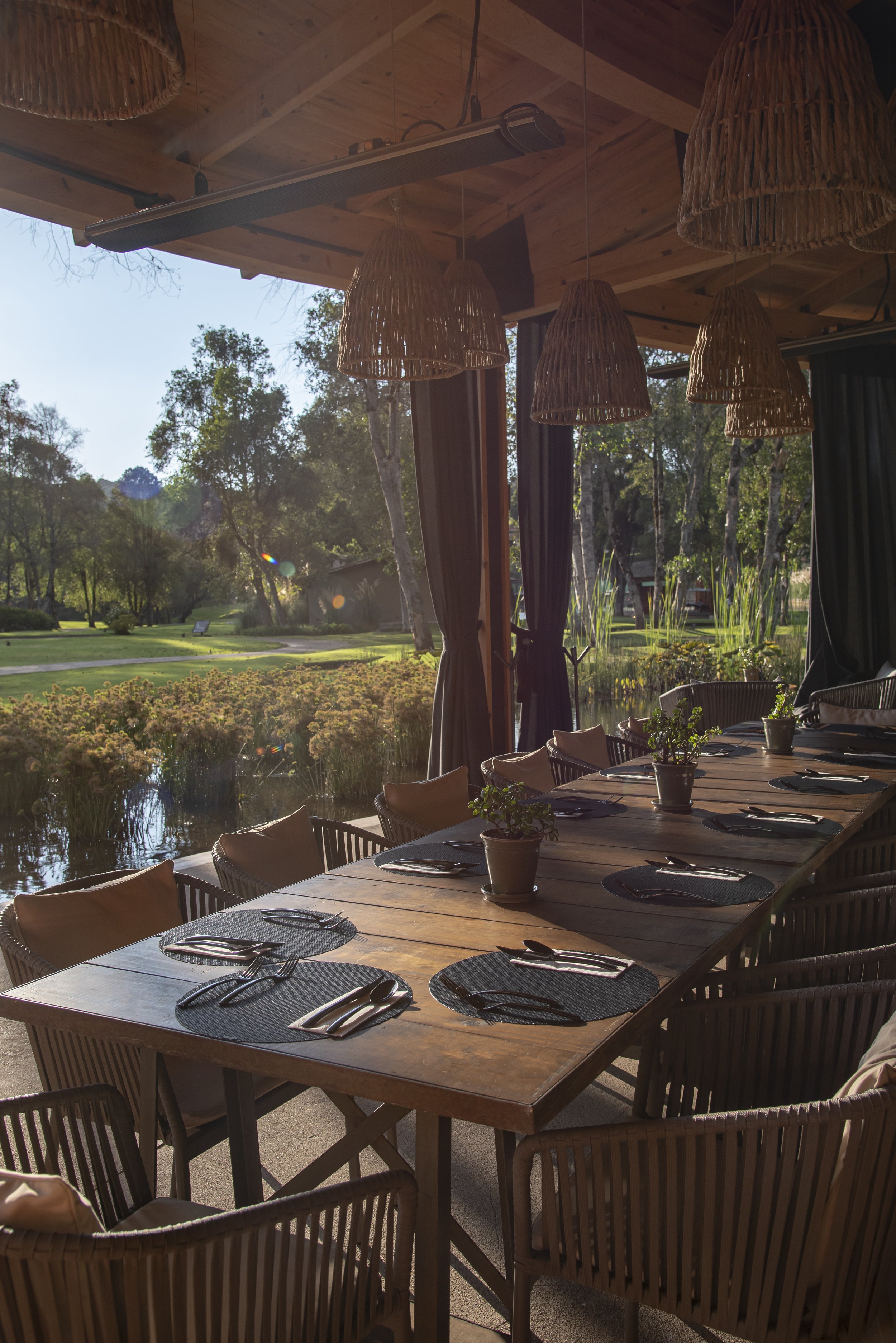 A wooden dining table set for a meal with black placemats, black napkins, silver utensils, and small potted plants, inside a room with large windows opening to a scenic outdoor view of a pond, trees, and a grassy area, with hanging wicker light fixtu