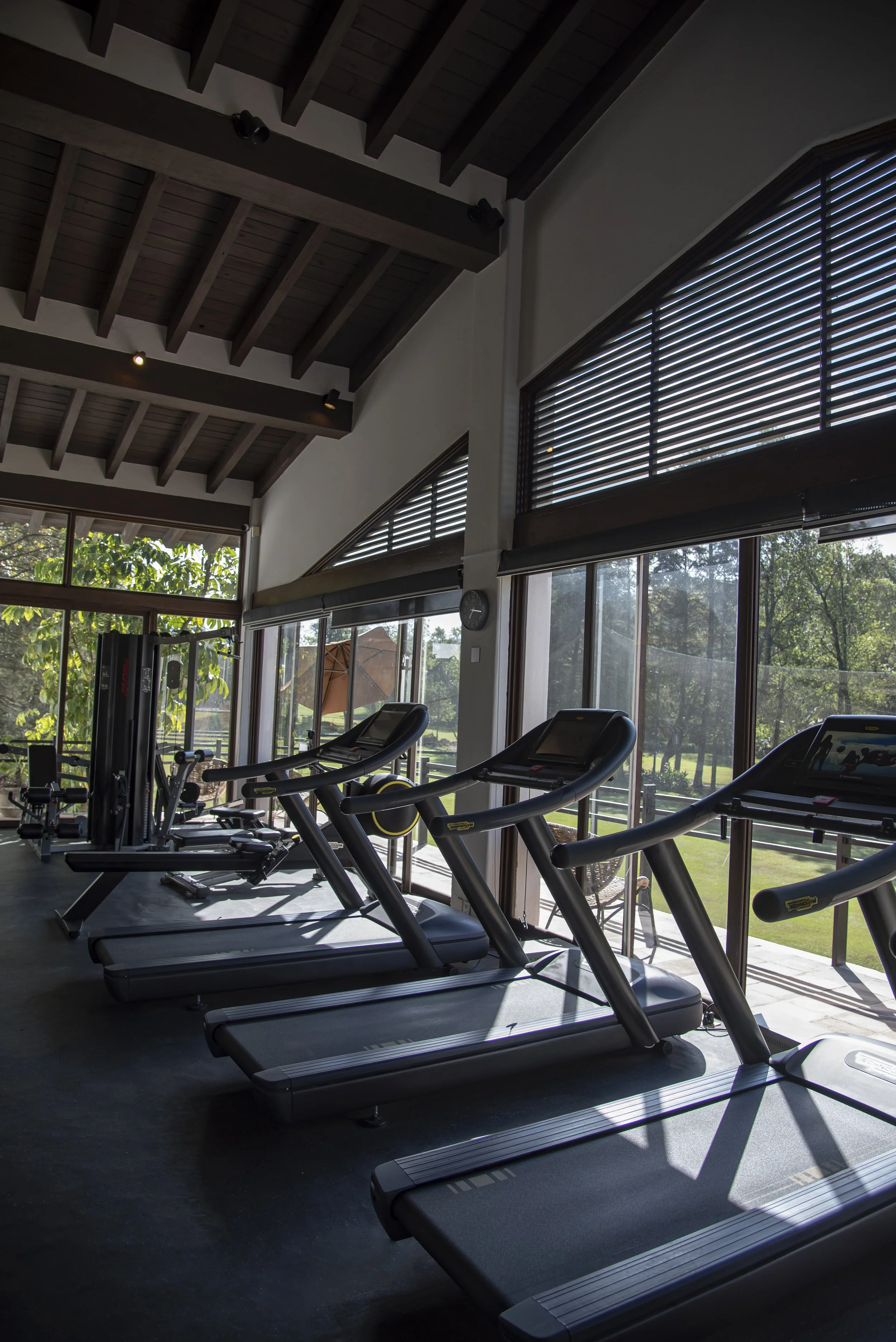 Interior of a gym with treadmills positioned near large windows with blinds, showing a view of green trees and outdoor patio area with chairs and an umbrella.