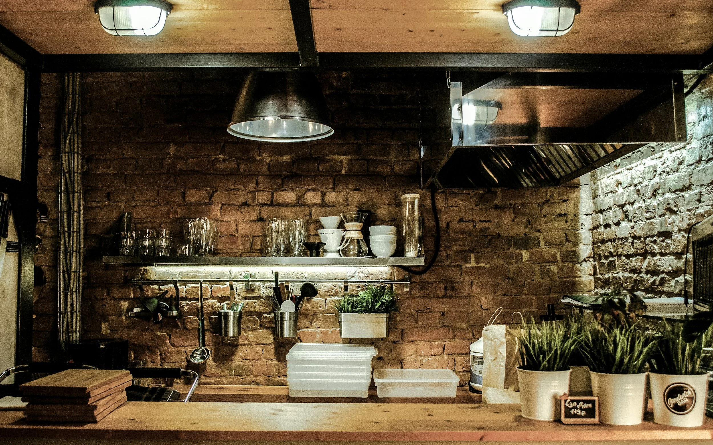 A cozy kitchen with an exposed brick wall, wooden countertop, and stainless steel range hood. Shelves hold glasses, bowls, and kitchen utensils. Potted plants decorate the counter.