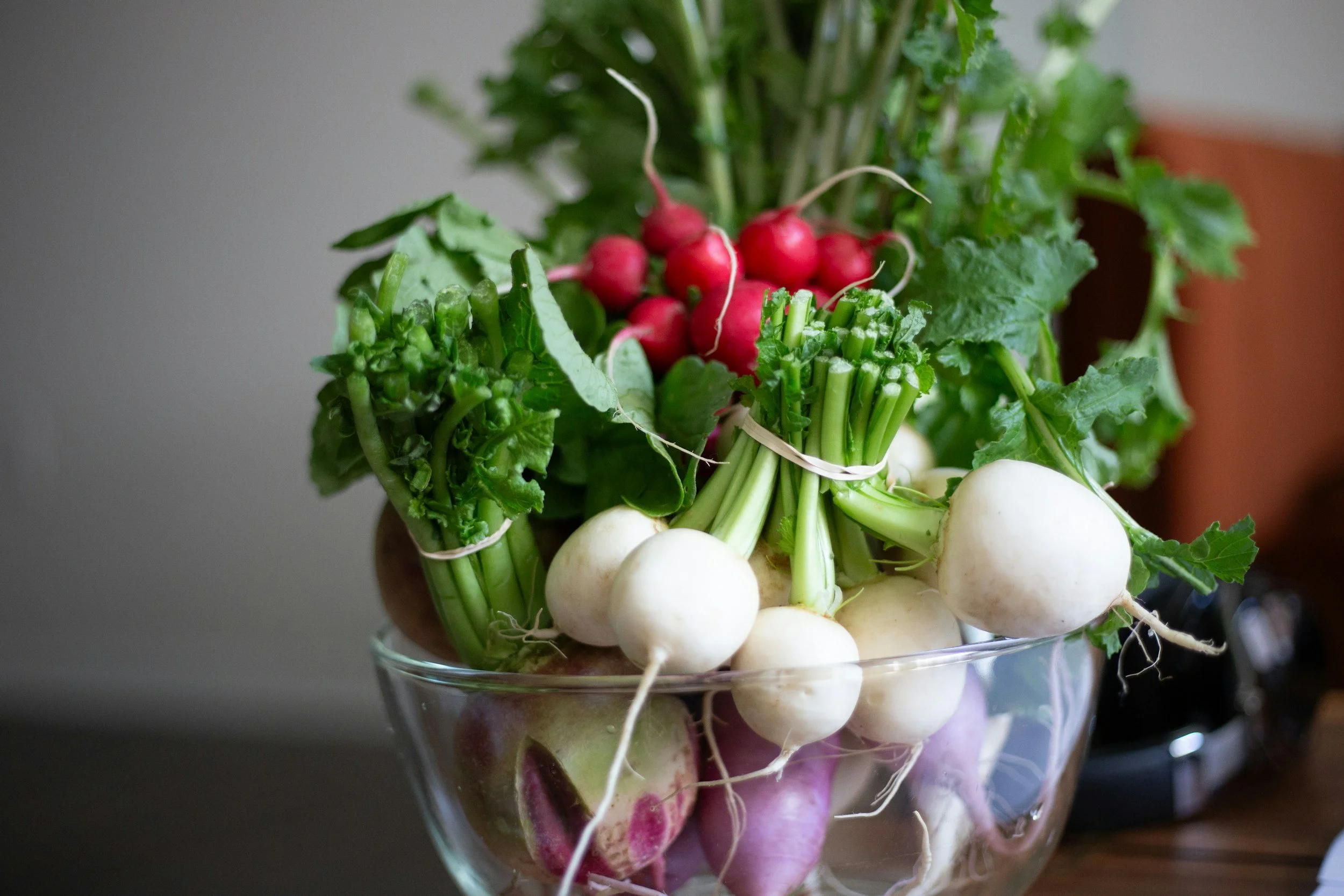 A glass bowl filled with assorted fresh vegetables, including radishes, turnips, and leafy greens.