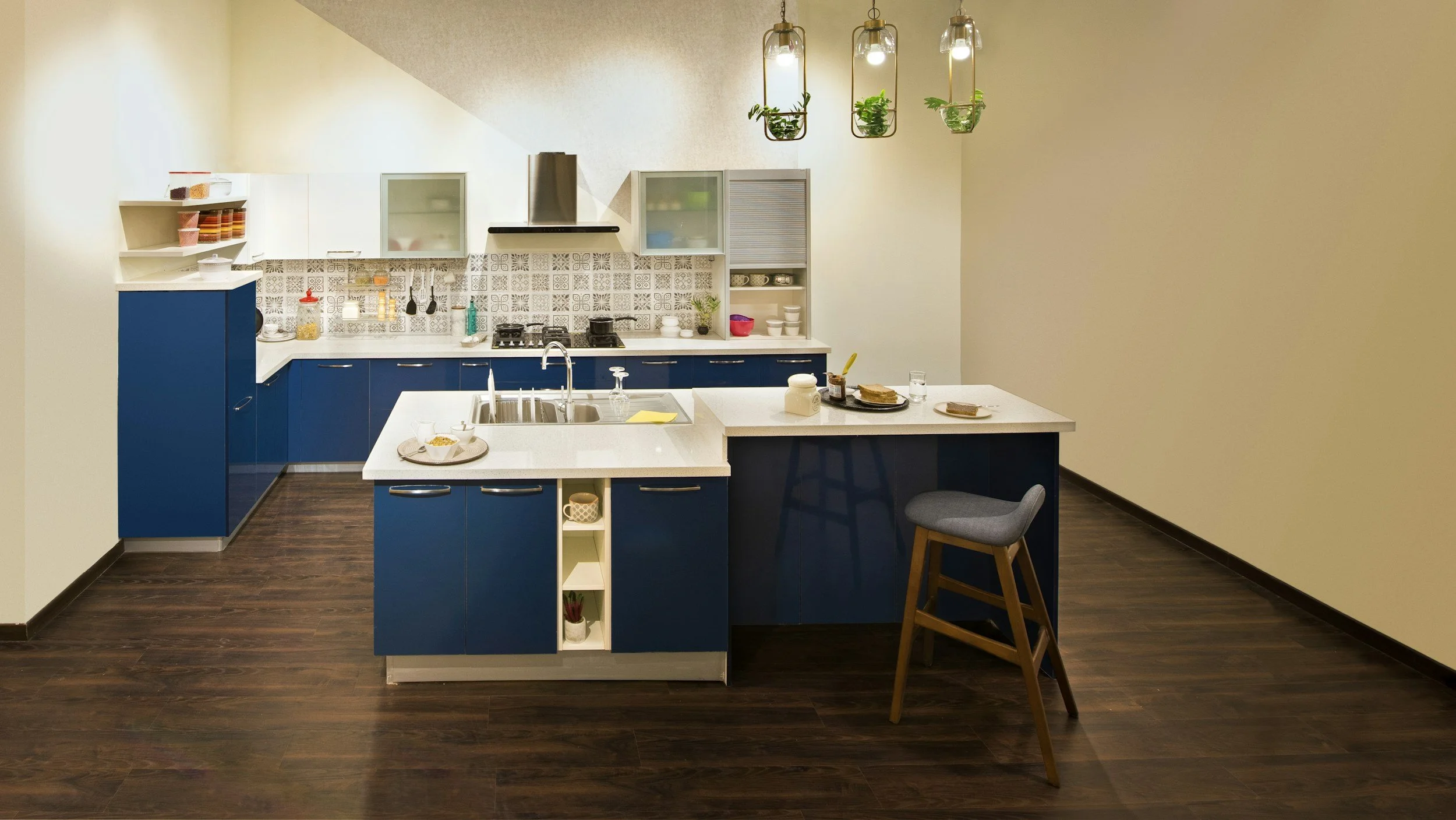 Modern kitchen with blue cabinets, white countertops, and a wooden floor. There is a kitchen island with a bar stool, a sink, and some dishes. The background wall has patterned tiles, open shelves, and hanging jars with plants, lights, and kitchen essentials.