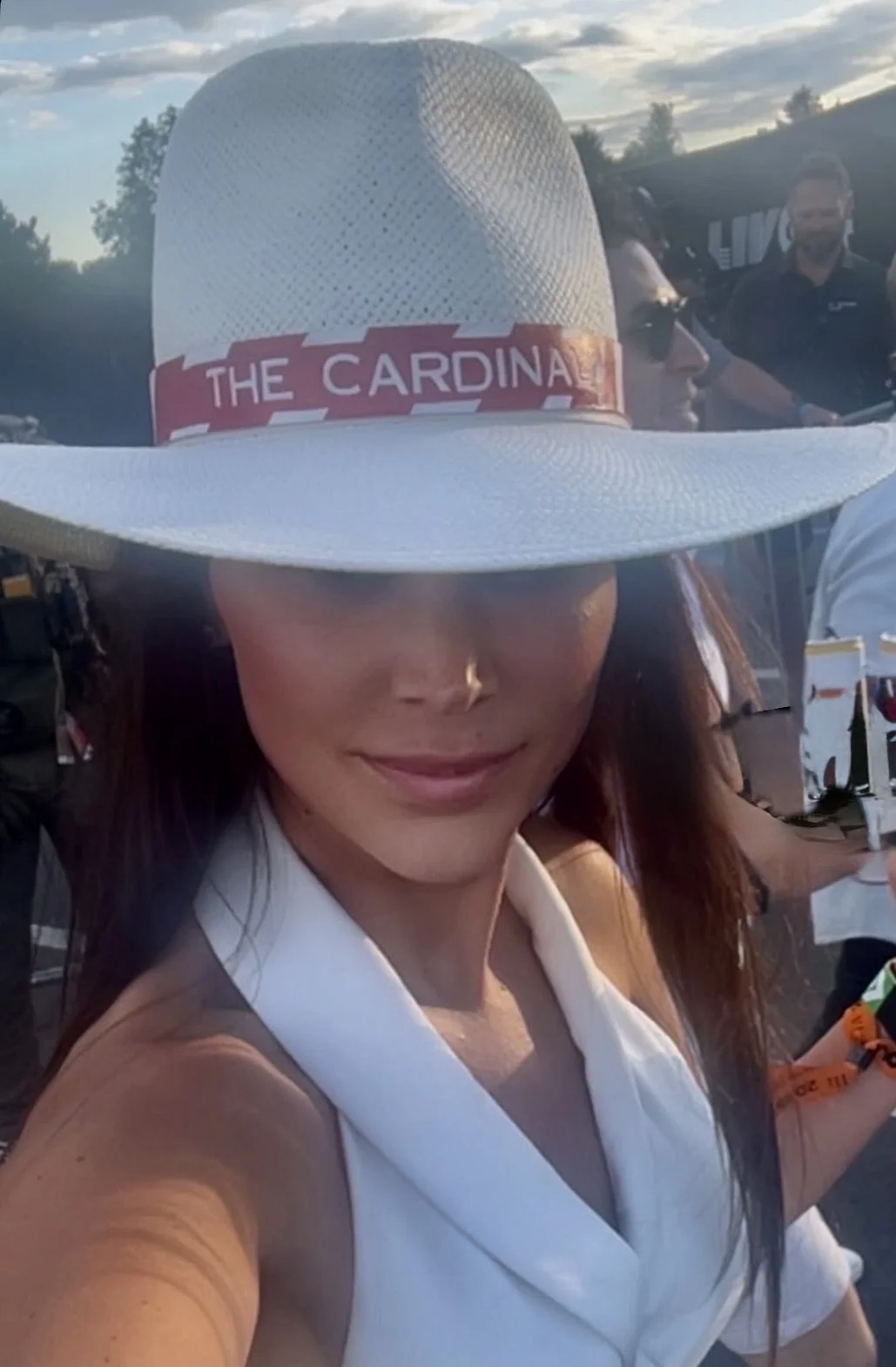 A woman taking a selfie, wearing a large white hat with a red band that says 'The Cardinal,' and a white sleeveless outfit, outdoors during sunset with a partly cloudy sky and other people in the background.