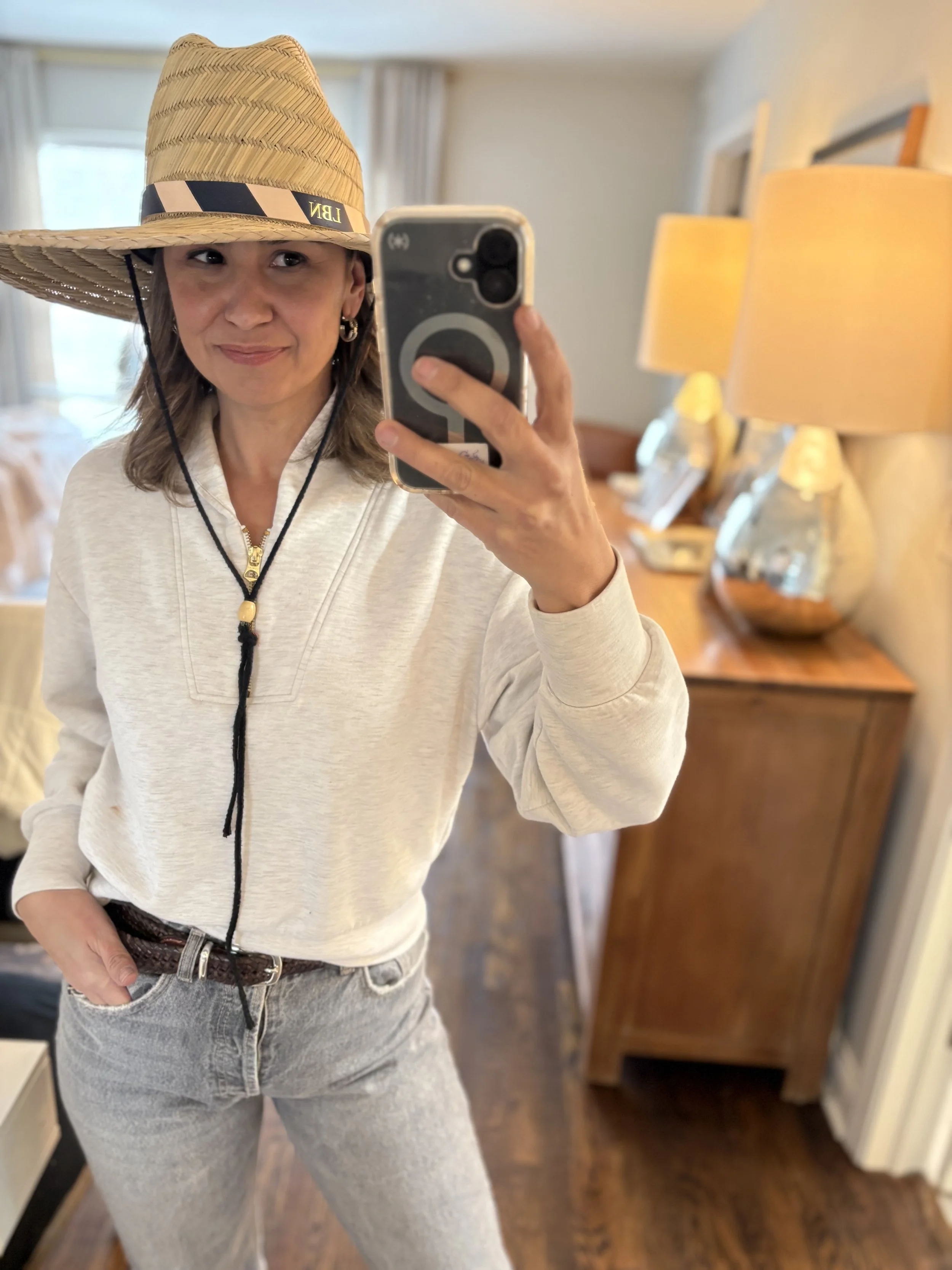 Woman taking a mirror selfie wearing a wide-brimmed straw hat, a cream-colored sweatshirt, and light gray jeans in a warmly lit room with wooden furniture and lamps.