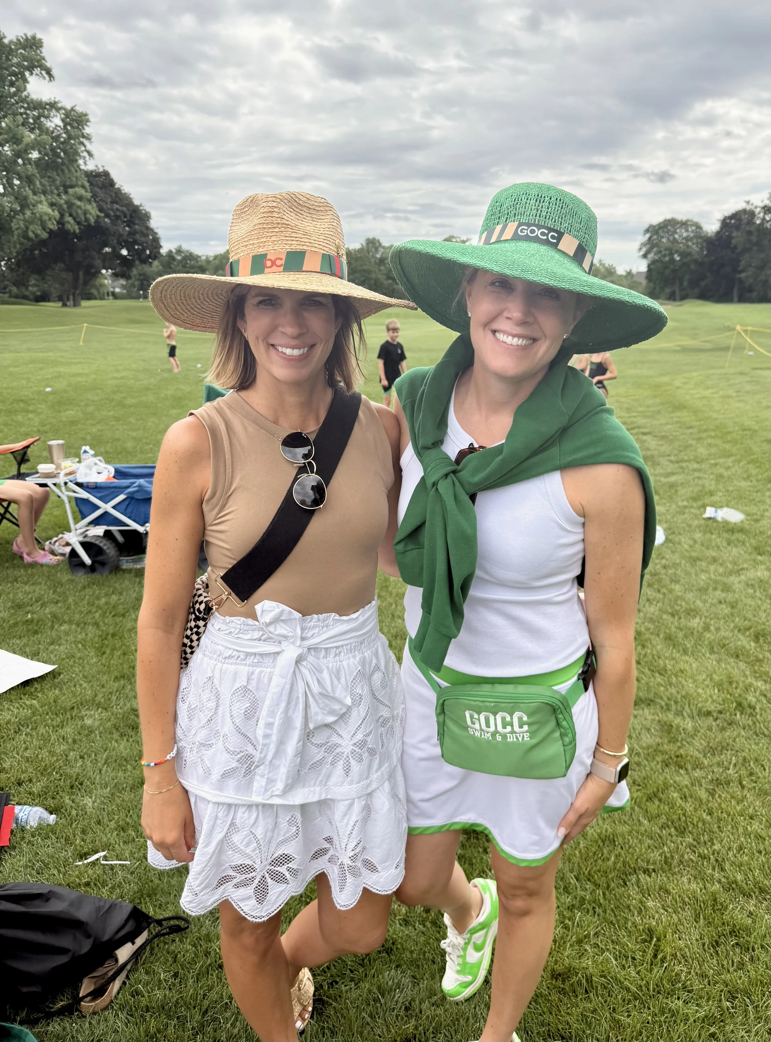 Two women standing outdoors on a grassy field, smiling, wearing large sun hats, with a group of people and trees in the background.