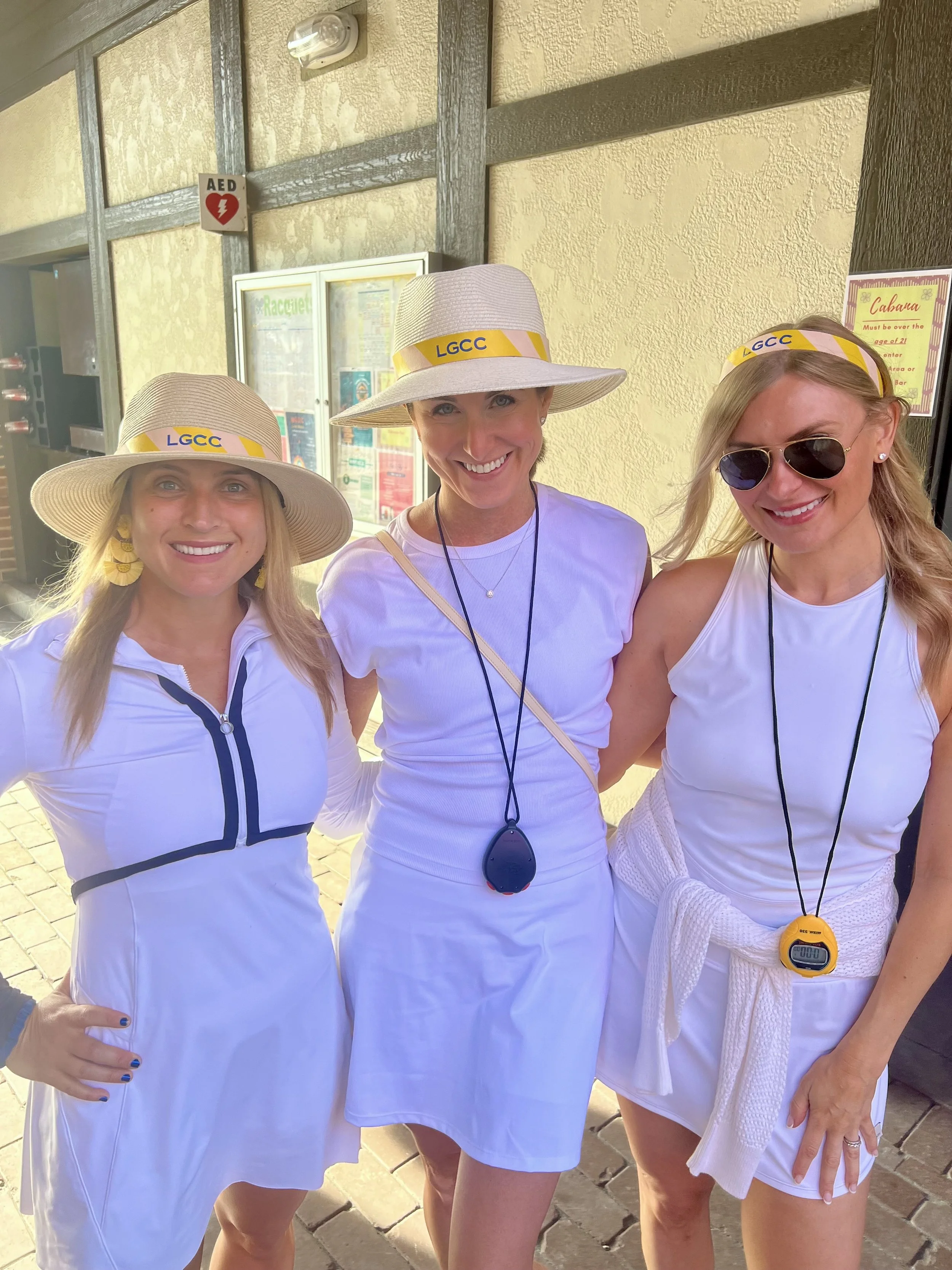Three women wearing white tennis outfits and wide-brimmed hats with yellow and purple 'LGCC' band, standing together outdoors, smiling, with one woman wearing sunglasses and another with a timer around her neck.