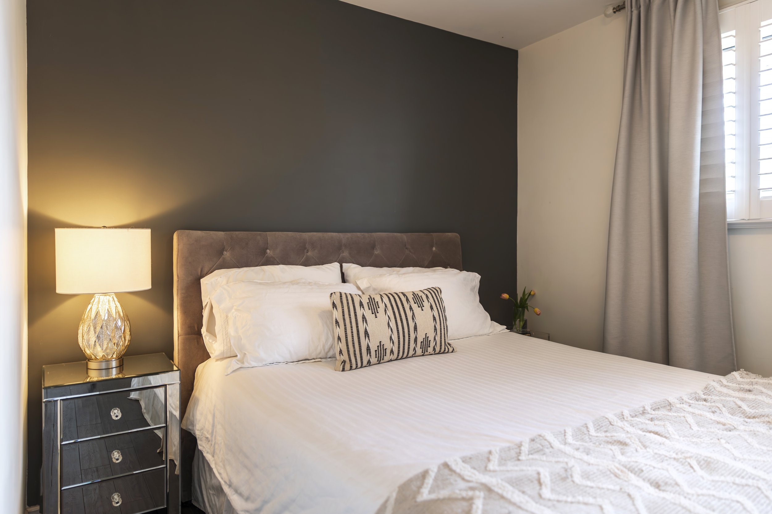 Modern bedroom with a bed featuring white linens and a patterned throw pillow, a mirrored nightstand with a lamp, and gray curtains.