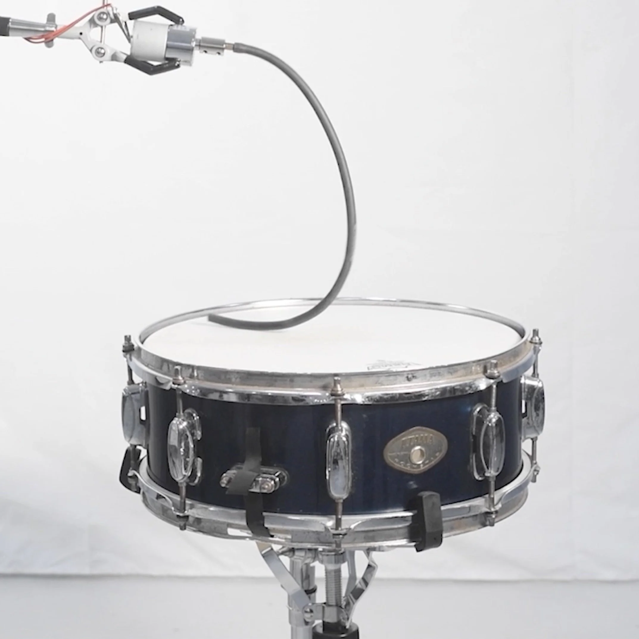 A  sound sculpture by artist, Bob Landstrom, featuring a snare drum with a motor positioned above it, mounted on a stand against a plain white background.