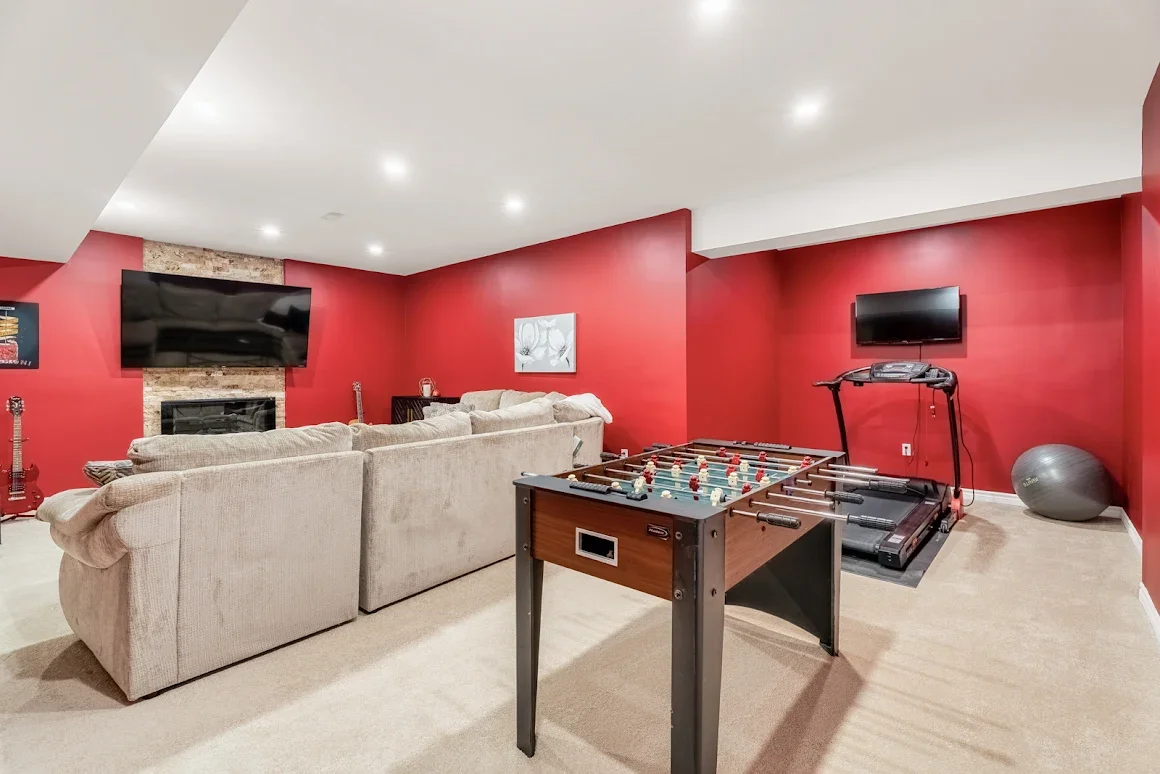Basement game room with red walls, beige sectional sofa, large flat-screen TV above a fireplace, foosball table, treadmill, exercise ball, and a painting on the wall.