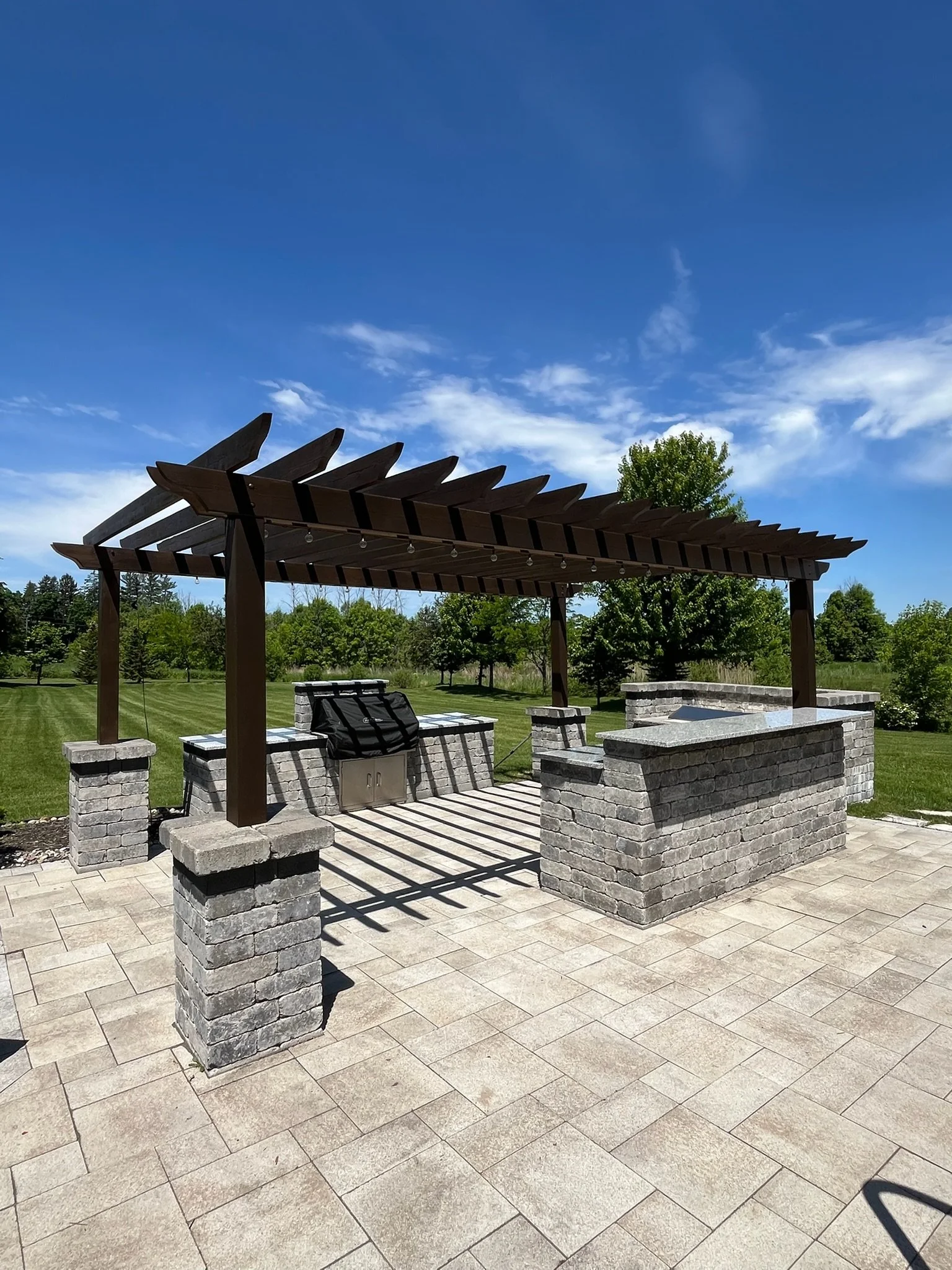 Outdoor stone patio with a brick barbecue grill under a wooden pergola, with a grassy yard and trees in the background.