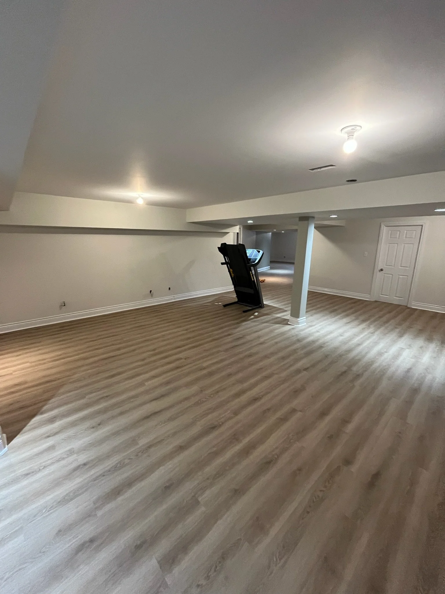 Empty basement room with light wood laminate flooring, white walls, and a white door. There are a few light bulbs in the ceiling and a treadmill leaning against a column.