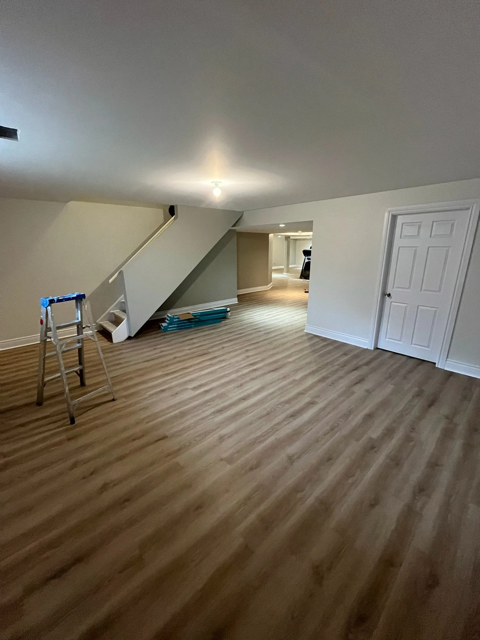 Empty room with a sloped ceiling, light-colored wood flooring, a small ladder, and a partially visible stairway. A door is on the right wall, and parts of a hallway or additional rooms are visible in the background, with minimal furnishings.