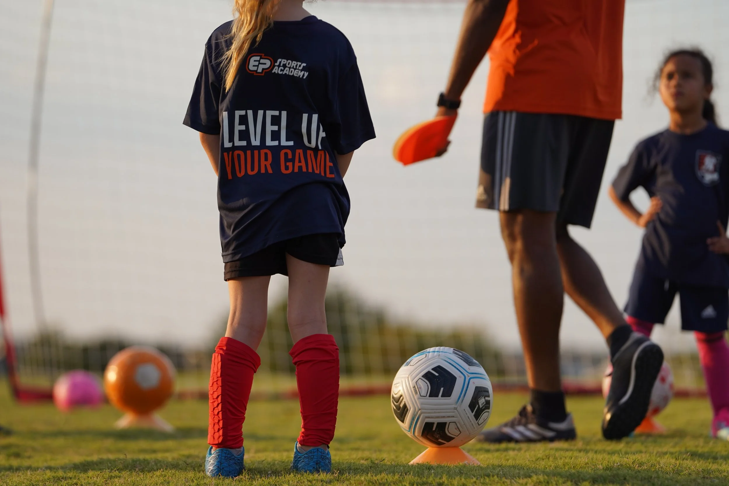 Children participating in a soccer practice on a field with orange cones, a coach observing, and soccer balls, under a metal pavilion on a sunny day.
