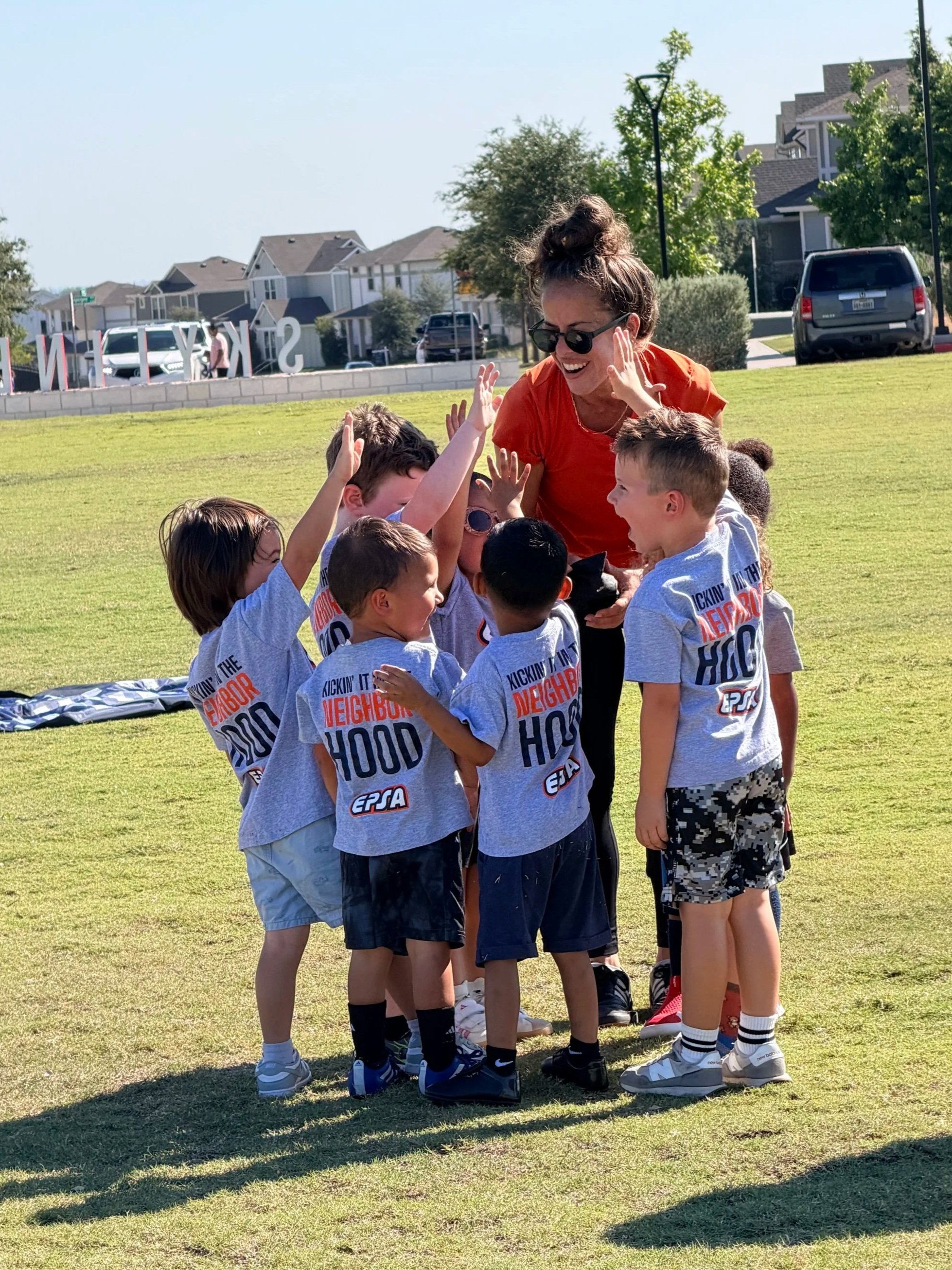 Four kids and one adult sitting on grass in a park, with two kids standing. The adult is wearing an orange shirt with 'SPORTS ACADEMY' printed on it. The kids are wearing orange vests and casual clothing, with three sitting and one standing, all engaging in a group activity. Trees, a cloudy sky, and park features are visible in the background.