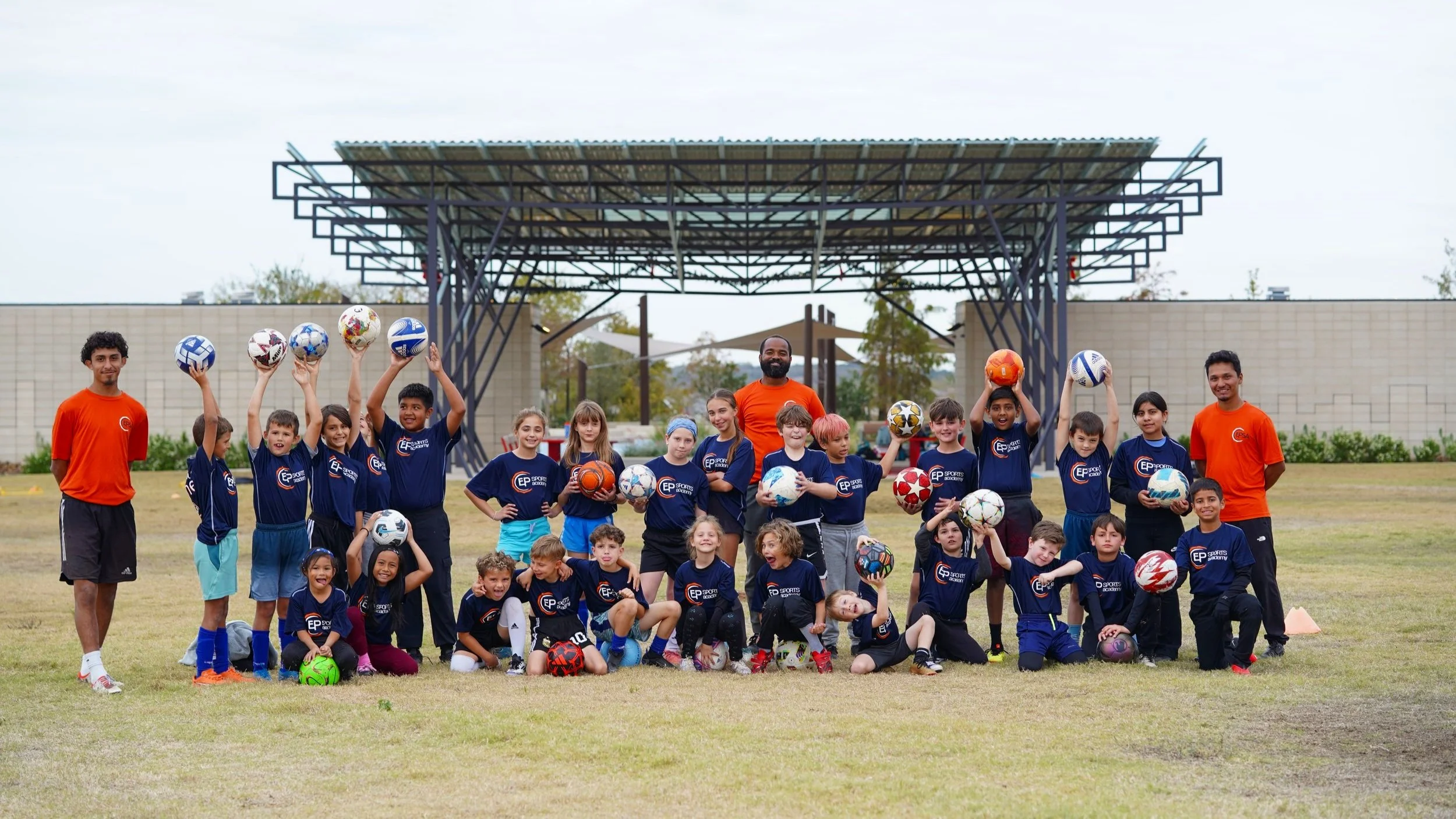 Group of children in blue shirts holding soccer balls, standing and kneeling on a grassy field with three adults in orange shirts.