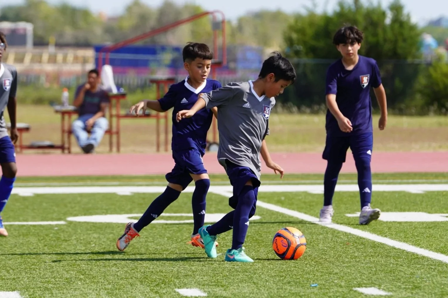 Group photo of children and coaches outdoors, standing on a ledge with a large tree and modern building in the background.