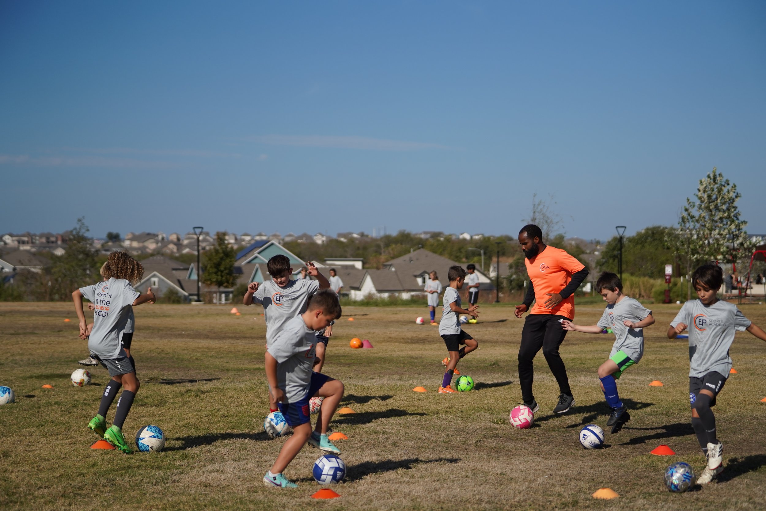 Children and a coach practicing soccer drills on a grassy field with cones and soccer balls in a residential area.