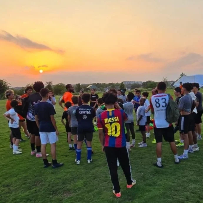 Child in a navy soccer uniform practices kicking a blue, white, and black soccer ball during an outdoor training session on a grassy field.