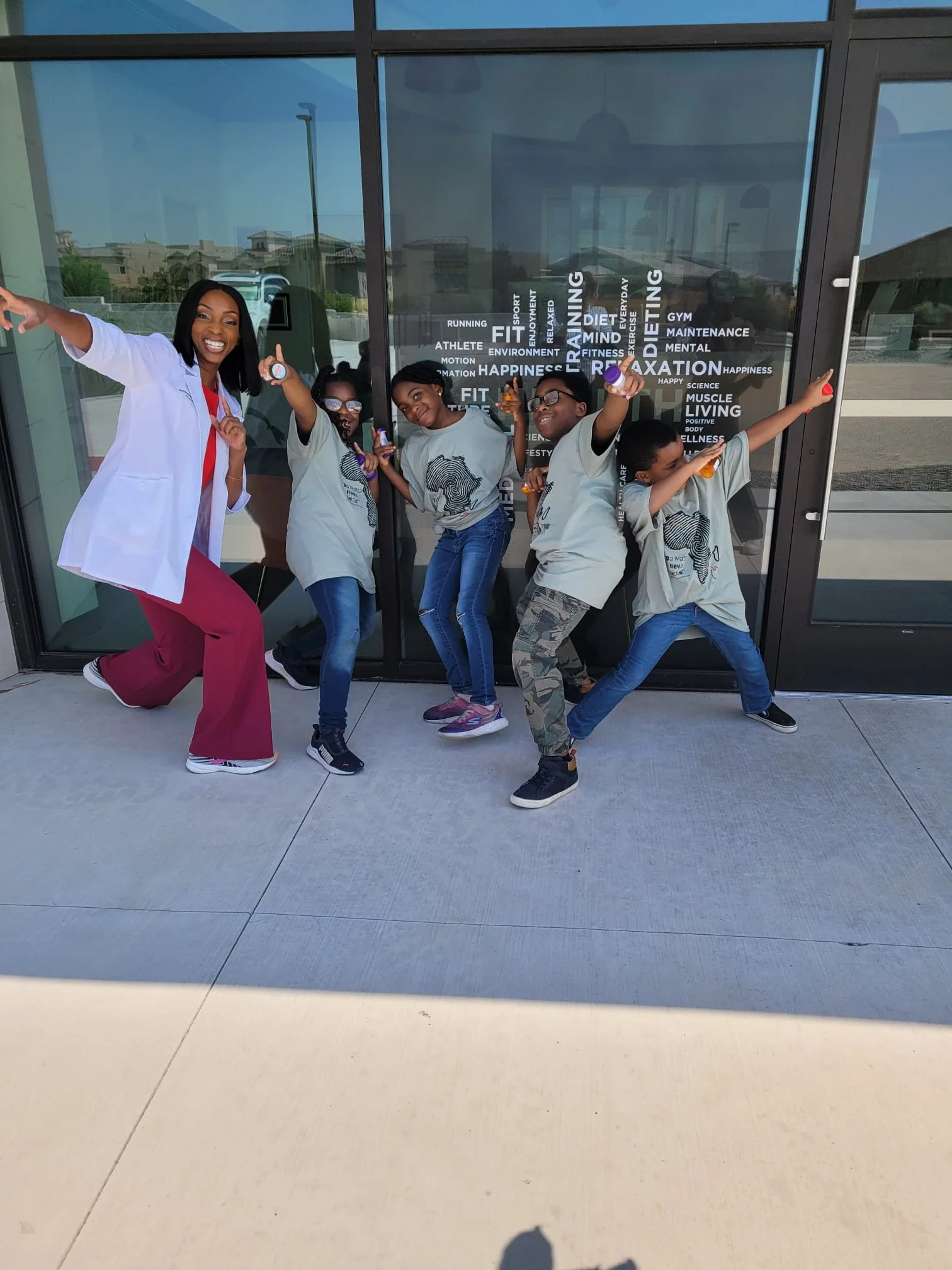 A woman in a white medical coat and five children in casual clothes posing playfully outside a building with large glass windows with words related to health, fitness, and happiness on the glass.