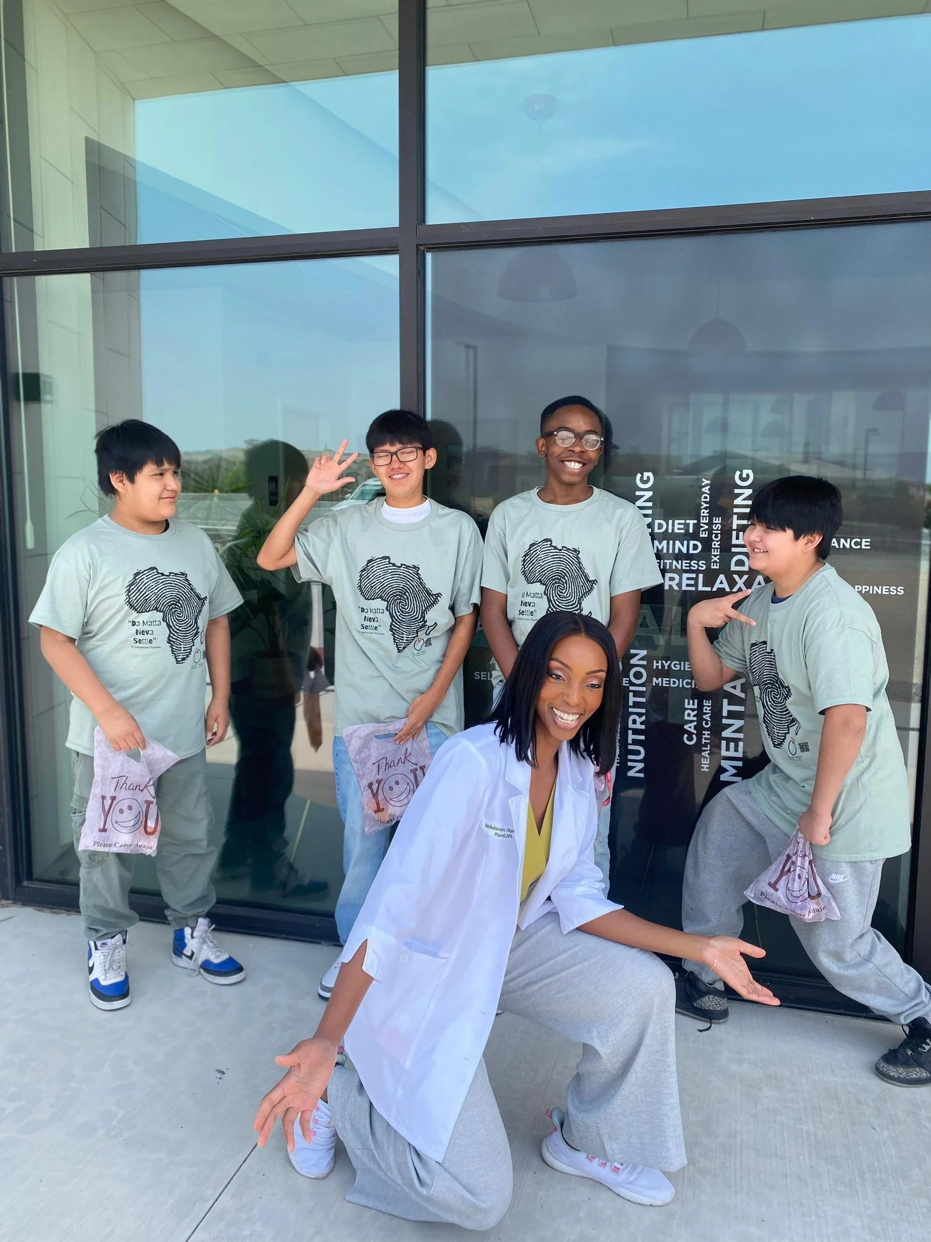 A smiling woman in a white coat kneeling in front of a group of boys wearing gray T-shirts with an African continent graphic, standing outside a modern building with large glass windows.