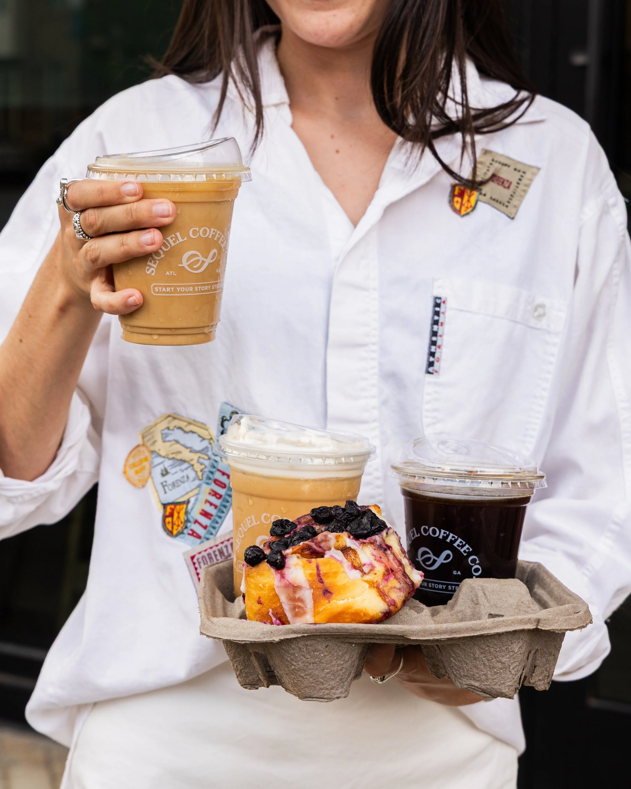 Customer holding cold lattes and coffee in to-go Sequel cups with a blueberry danish