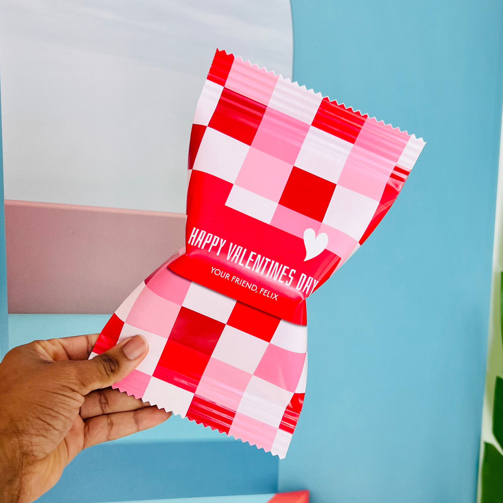 Hand holding a pink and red checkered Valentine’s Day wrapper with the text 'Happy Valentines Day' and 'Your Friend, Felix' against a colorful background.