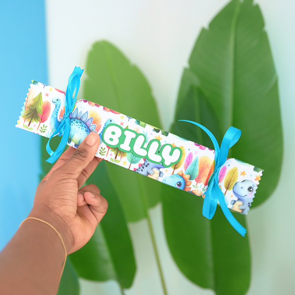Person holding a decorated chocolate wafer bar wrapped in colorful themed paper with the name 'Bilco' in green letters, tied with blue ribbons, in front of large green leaves.