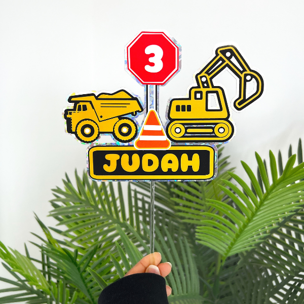 Decorative construction-themed sign with a yellow dump truck, yellow excavator, octagonal red stop sign with the number 3, orange and white traffic cone, and a black and yellow sign that says "JUDOH." Green palm leaves are at the bottom.