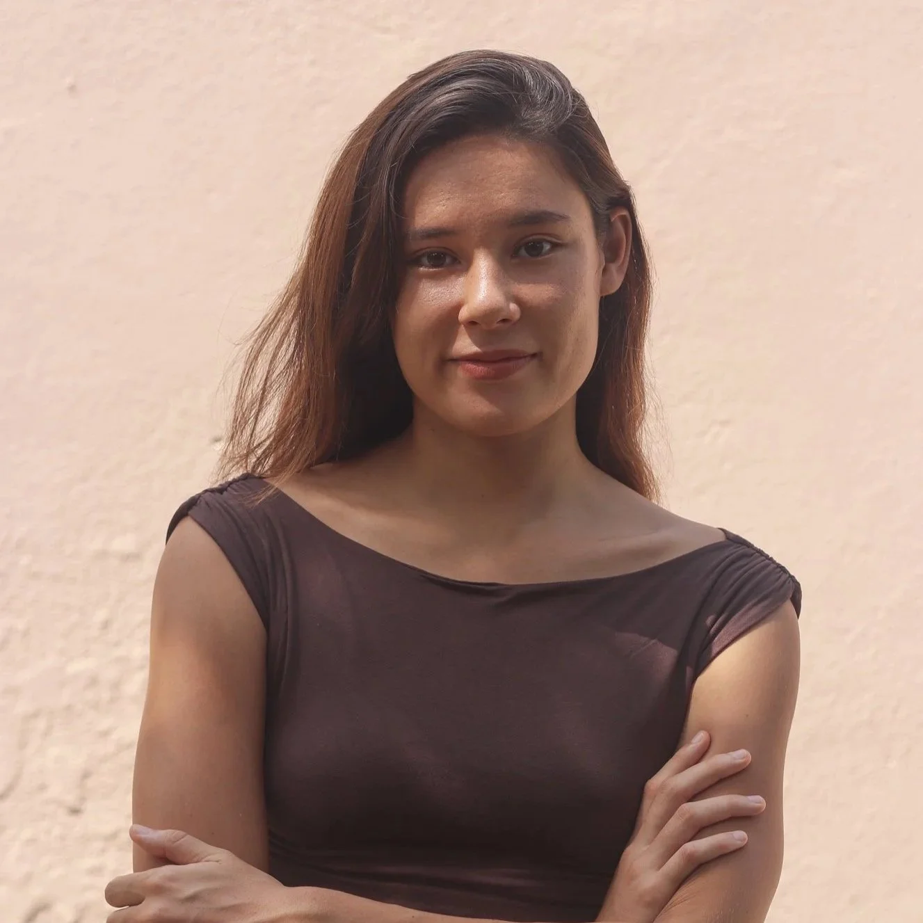 A young woman with long brown hair, wearing a dark sleeveless top, standing against a beige textured wall, with her arms crossed.