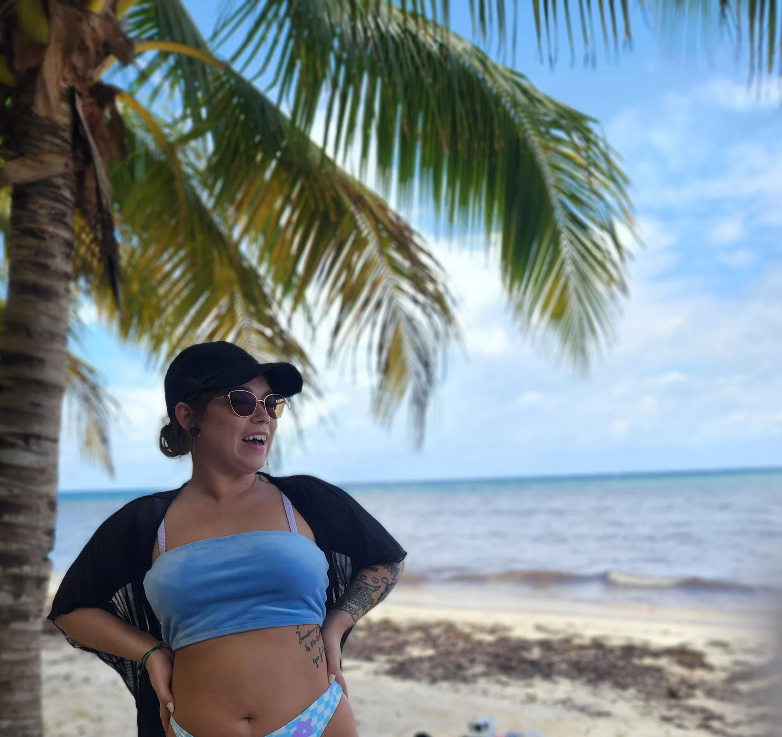 Person in a bikini and sunglasses under a palm tree at the beach.