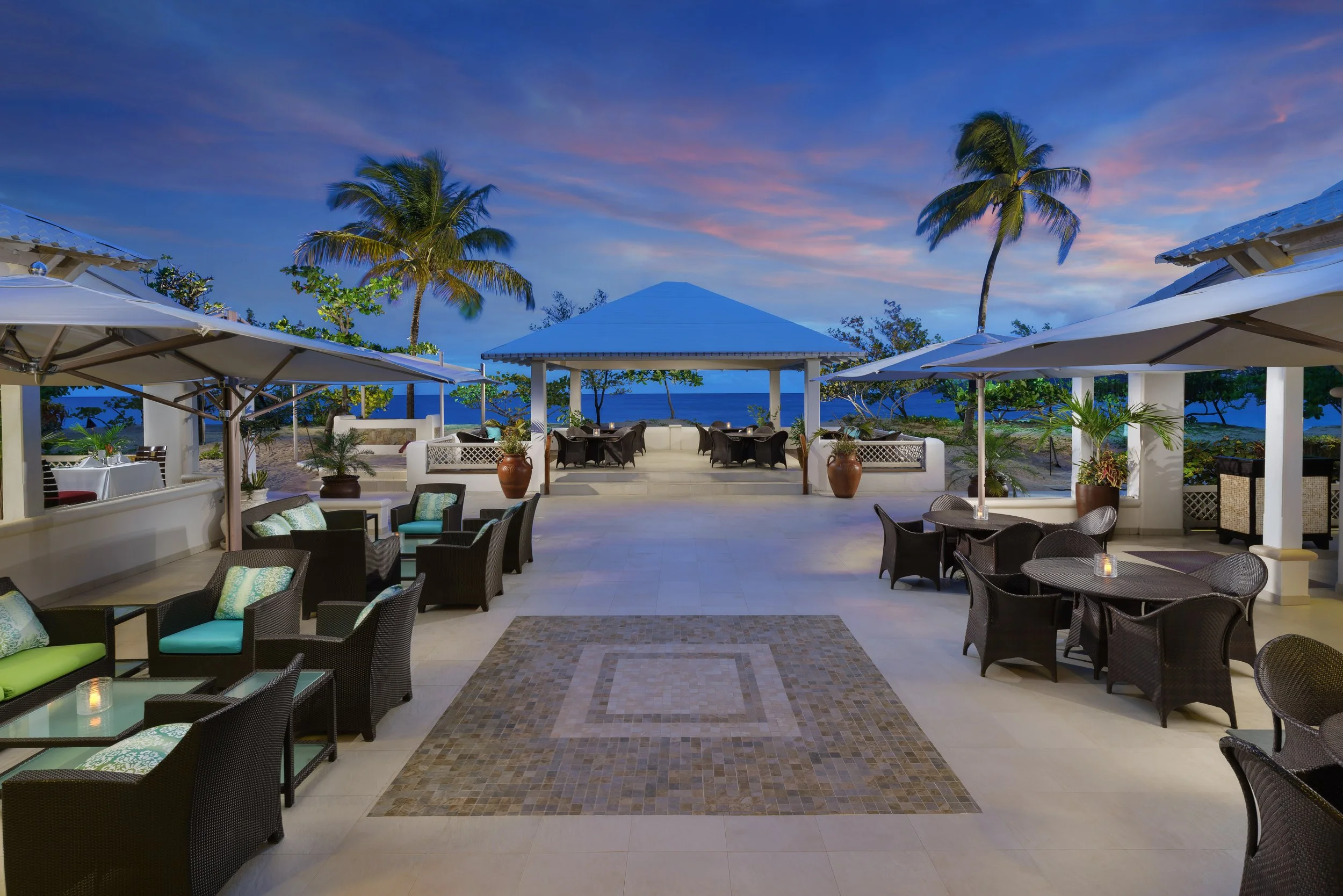 Outdoor restaurant patio with wicker furniture, palm trees, and ocean view at sunset