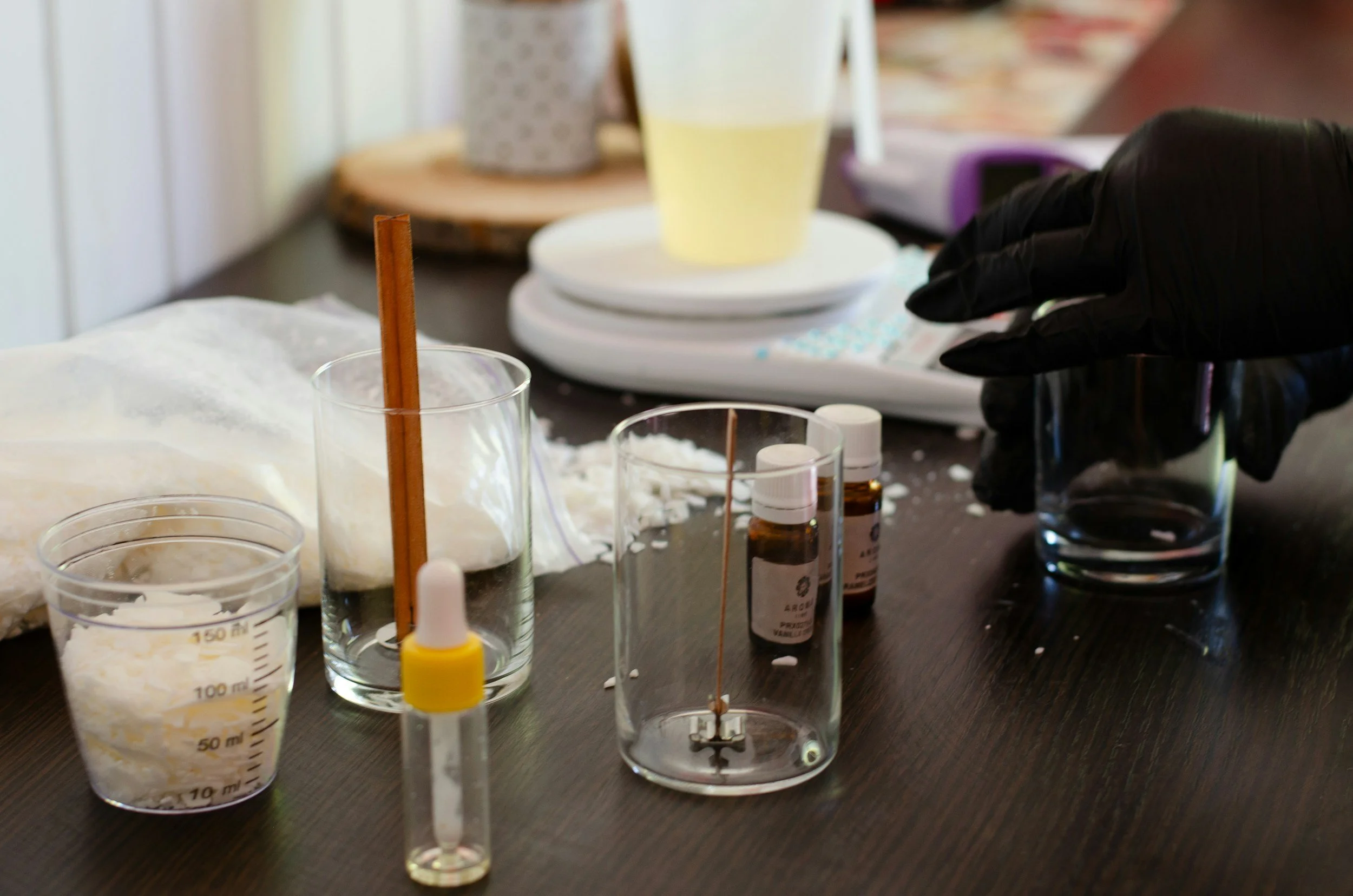 A medical or laboratory setup with various glass beakers, small bottles, and medical supplies on a dark wooden table, with a gloved hand reaching for a glass.