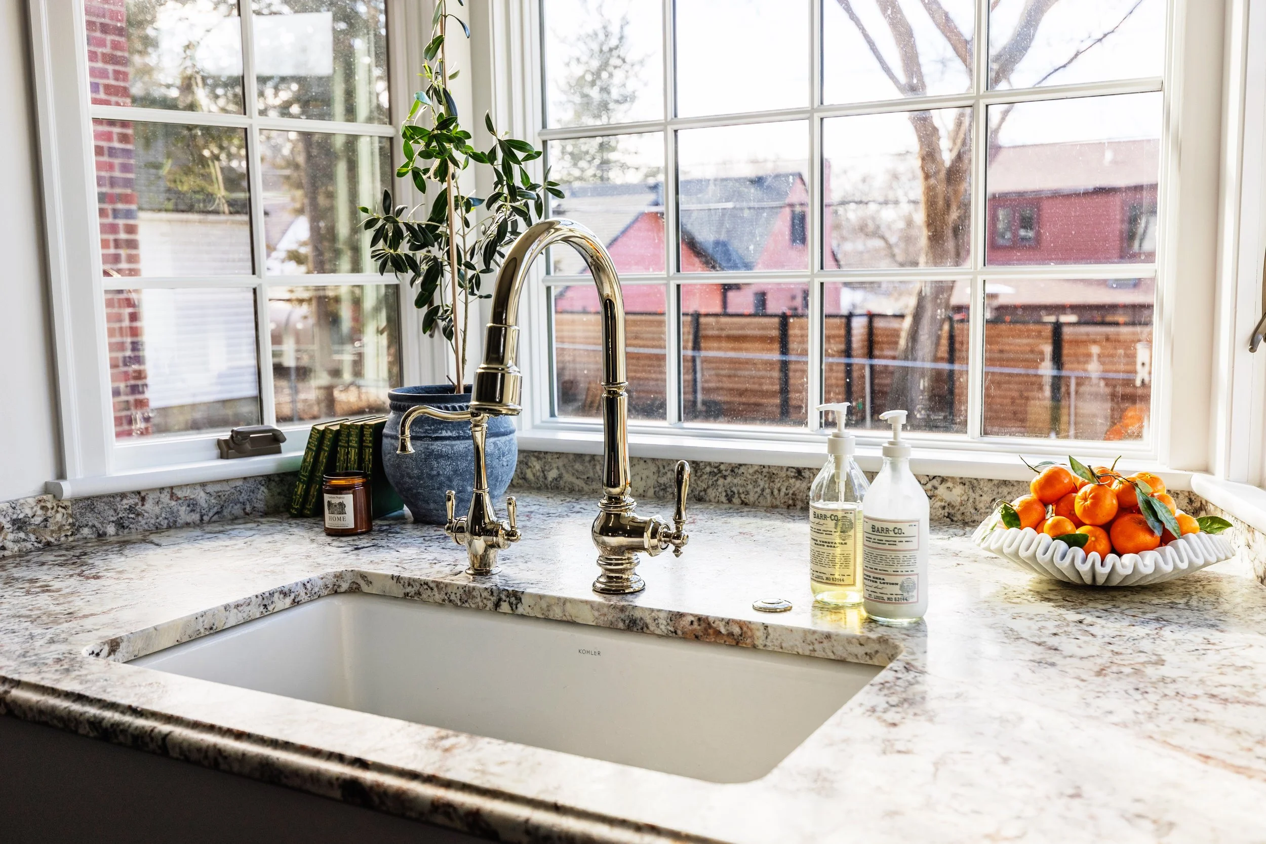 Kitchen sink with granite countertop, two soap dispensers, a bowl of oranges, and a potted plant near a large window showing an outdoor backyard scene.