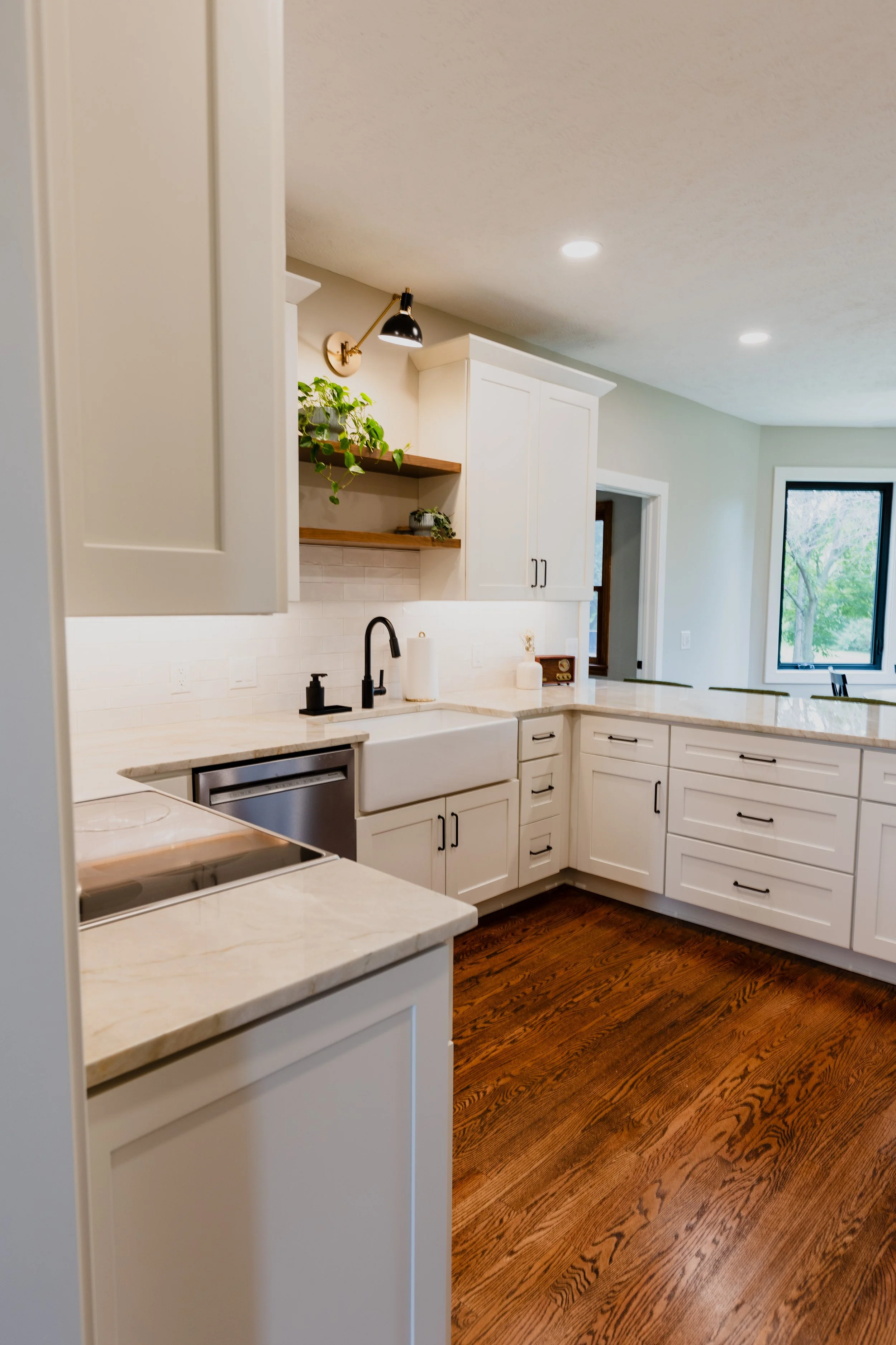 Modern kitchen with white cabinets, marble countertops, and hardwood floors, featuring a farmhouse sink and a window with a tree outside.