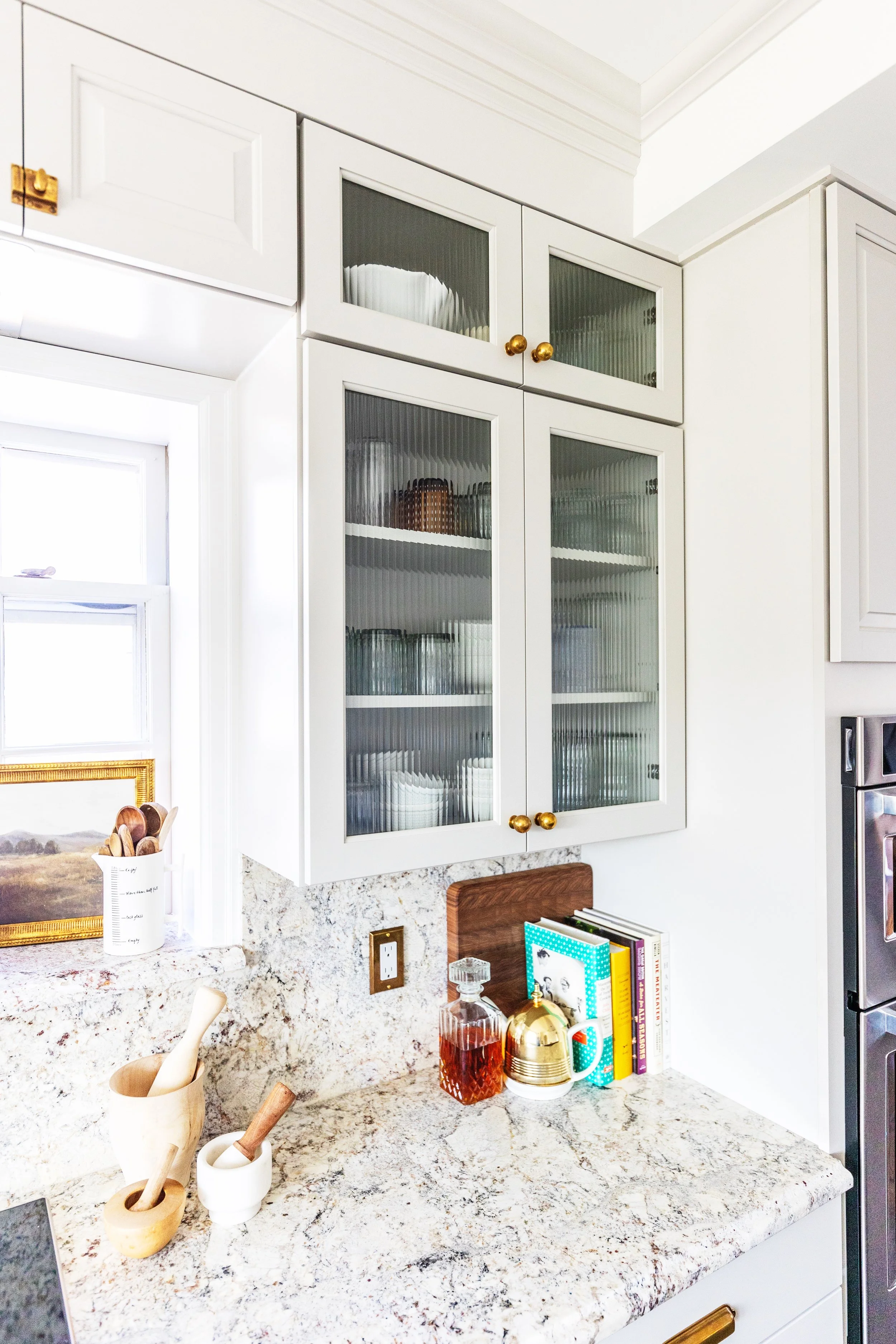 White kitchen with glass-front cabinets, granite countertop, and decorative items including a decanter, books, and mortar and pestles.