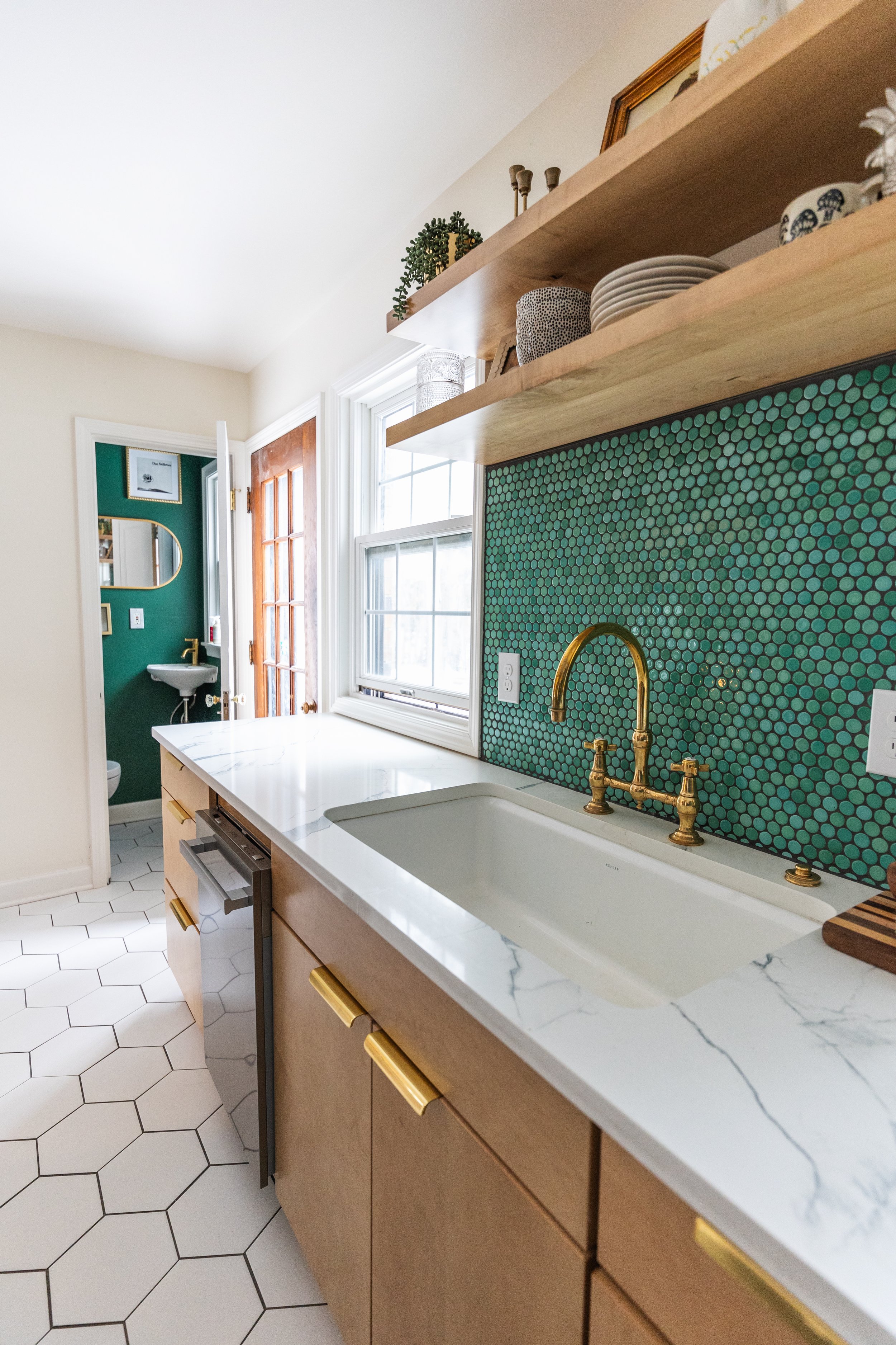 Kitchen with white marble countertop, gold hardware cabinets, green mosaic tile backsplash, brass faucet, and open wooden shelves with dishes and decor.
