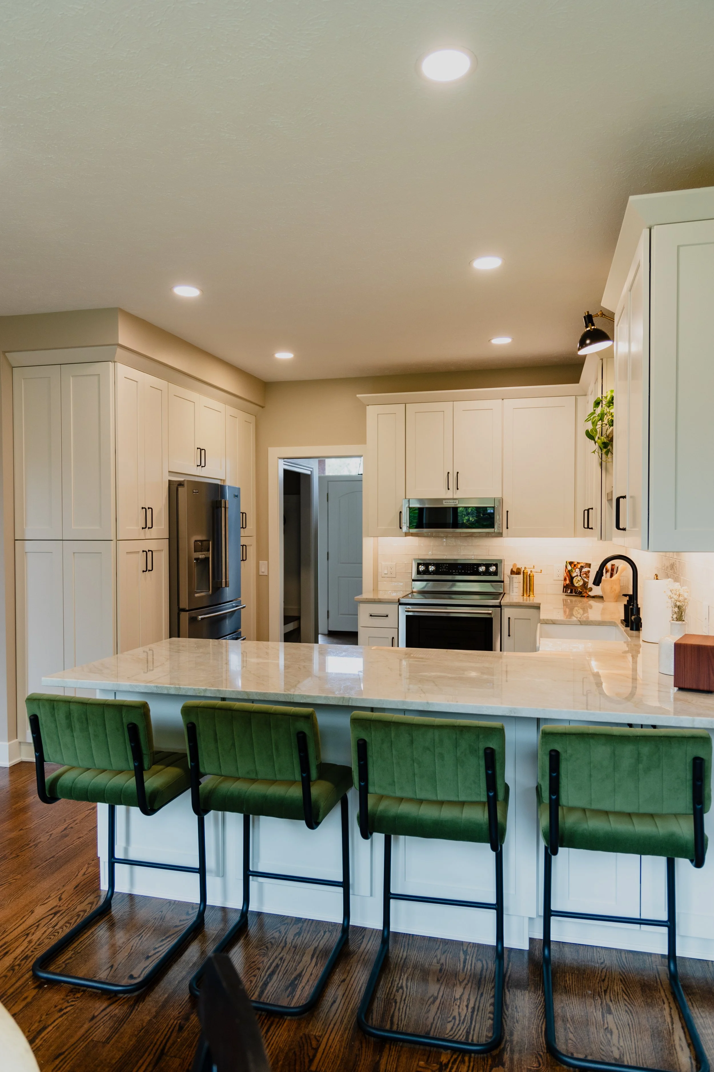 Modern kitchen with white cabinets, stainless steel appliances, marble island, green cushioned bar stools, and wooden flooring.