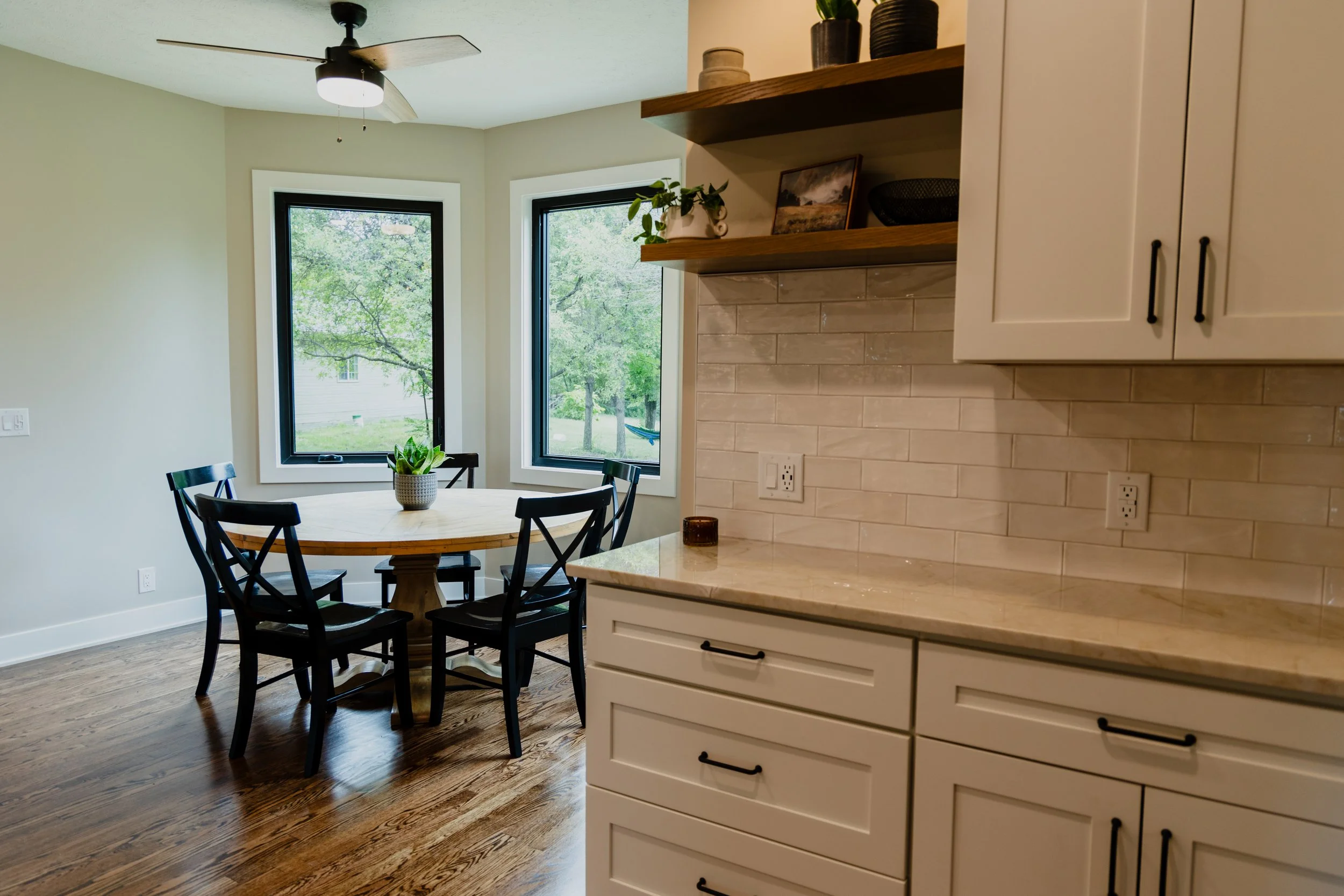 Kitchen with white cabinets, quartz countertop, and a round dining table with four black chairs near two large windows. 1935 Kitchen Remodel. Black Matte Hardware. White Cabinets. White Subway Backsplash. Eat in Kitchen.