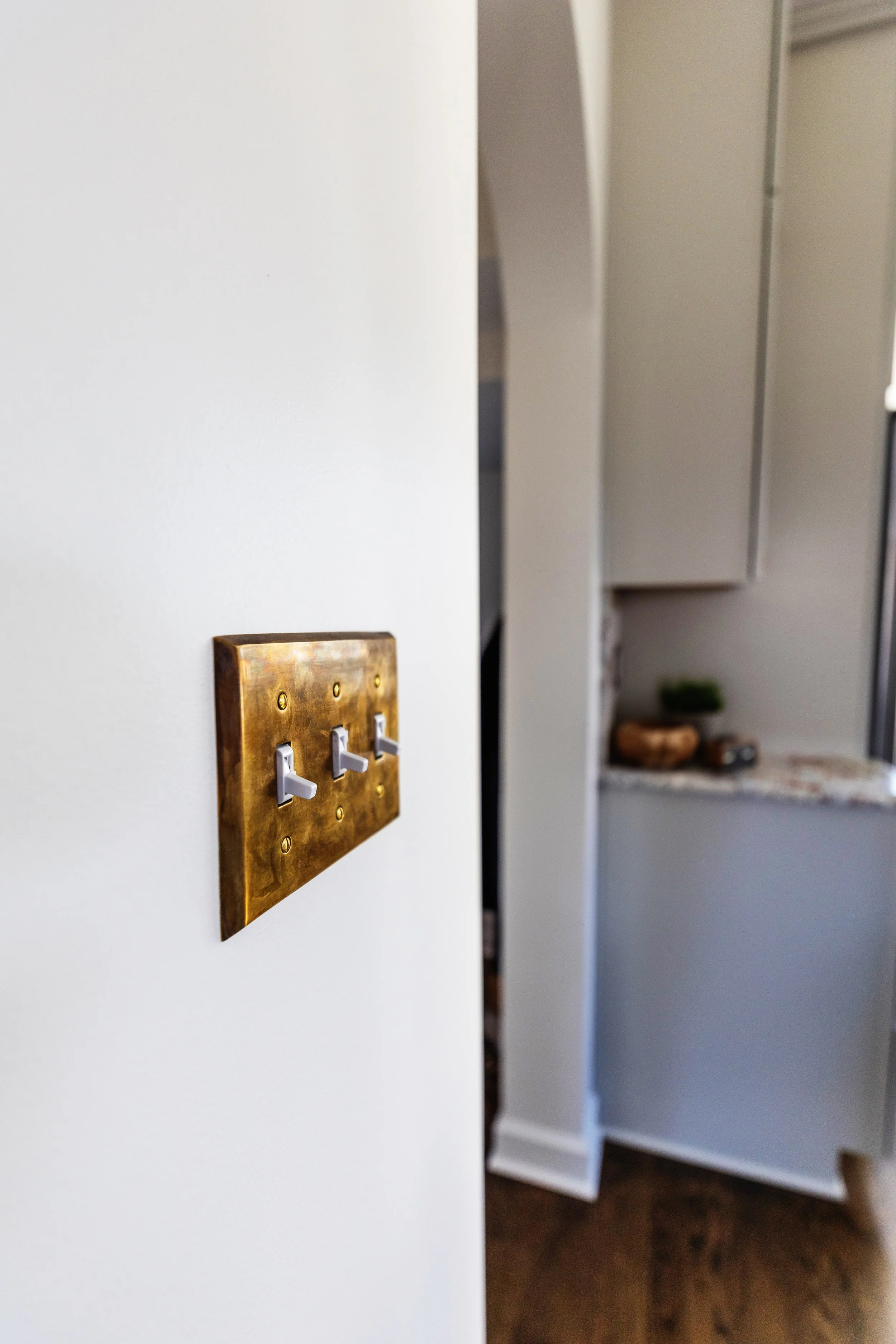 Close-up of a brass light switch plate with three white toggle switches on a white wall in a kitchen or living space.