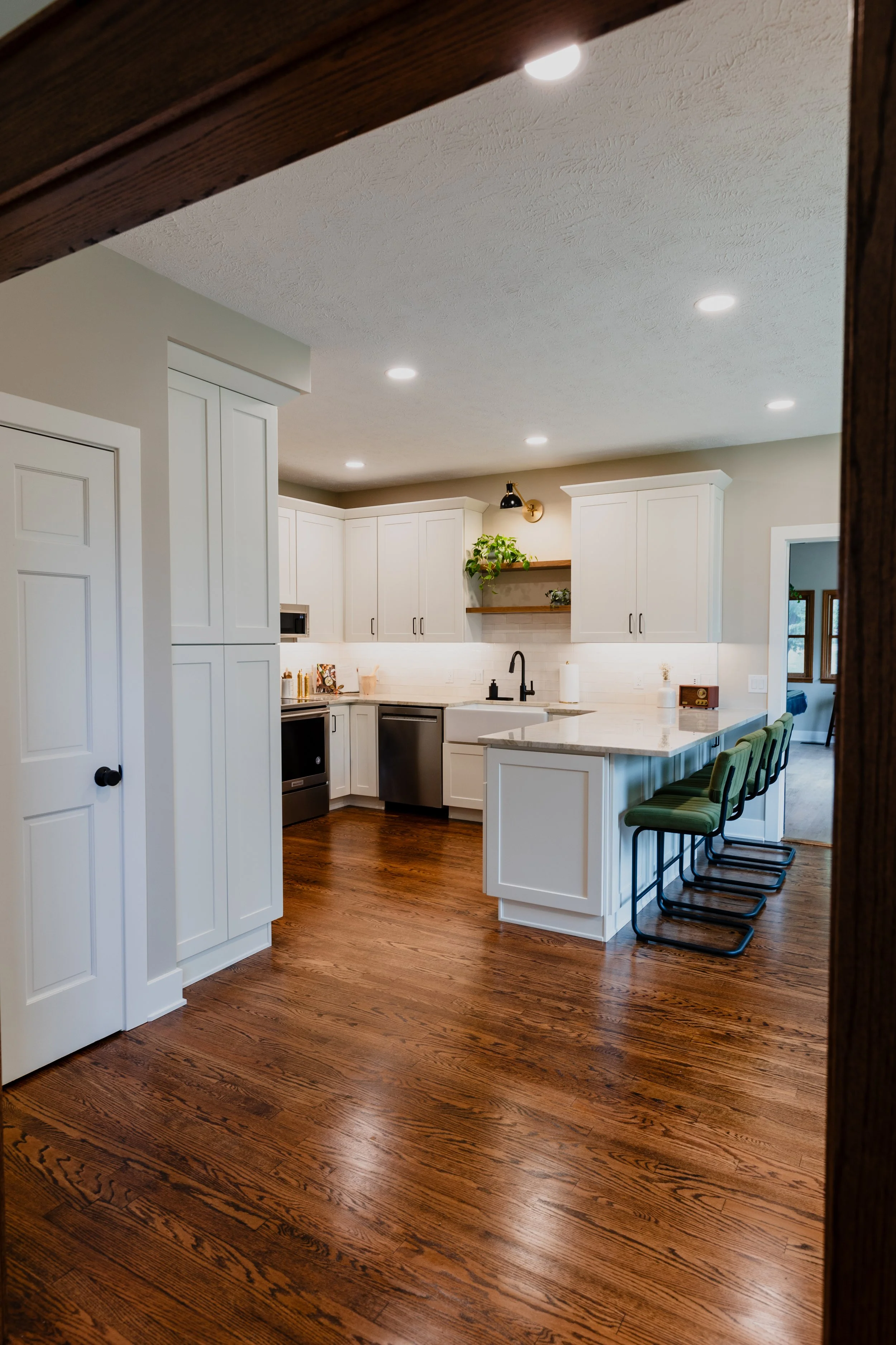 Modern kitchen with white cabinets, a marble countertop island with four green chairs, hardwood floors, and recessed ceiling lights.