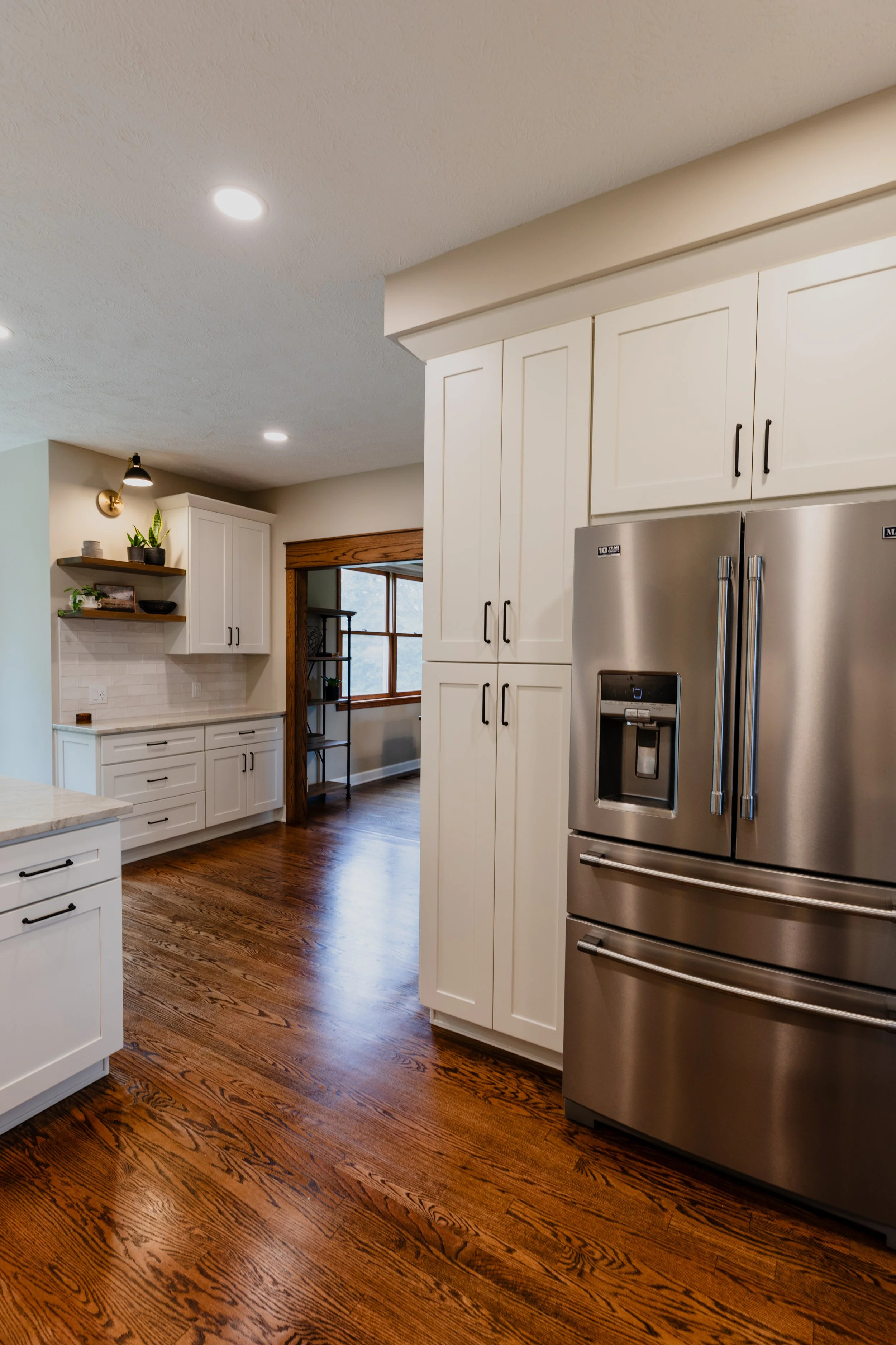 Modern kitchen with stainless steel refrigerator, white cabinets, wooden floor, and a sitting area with window.