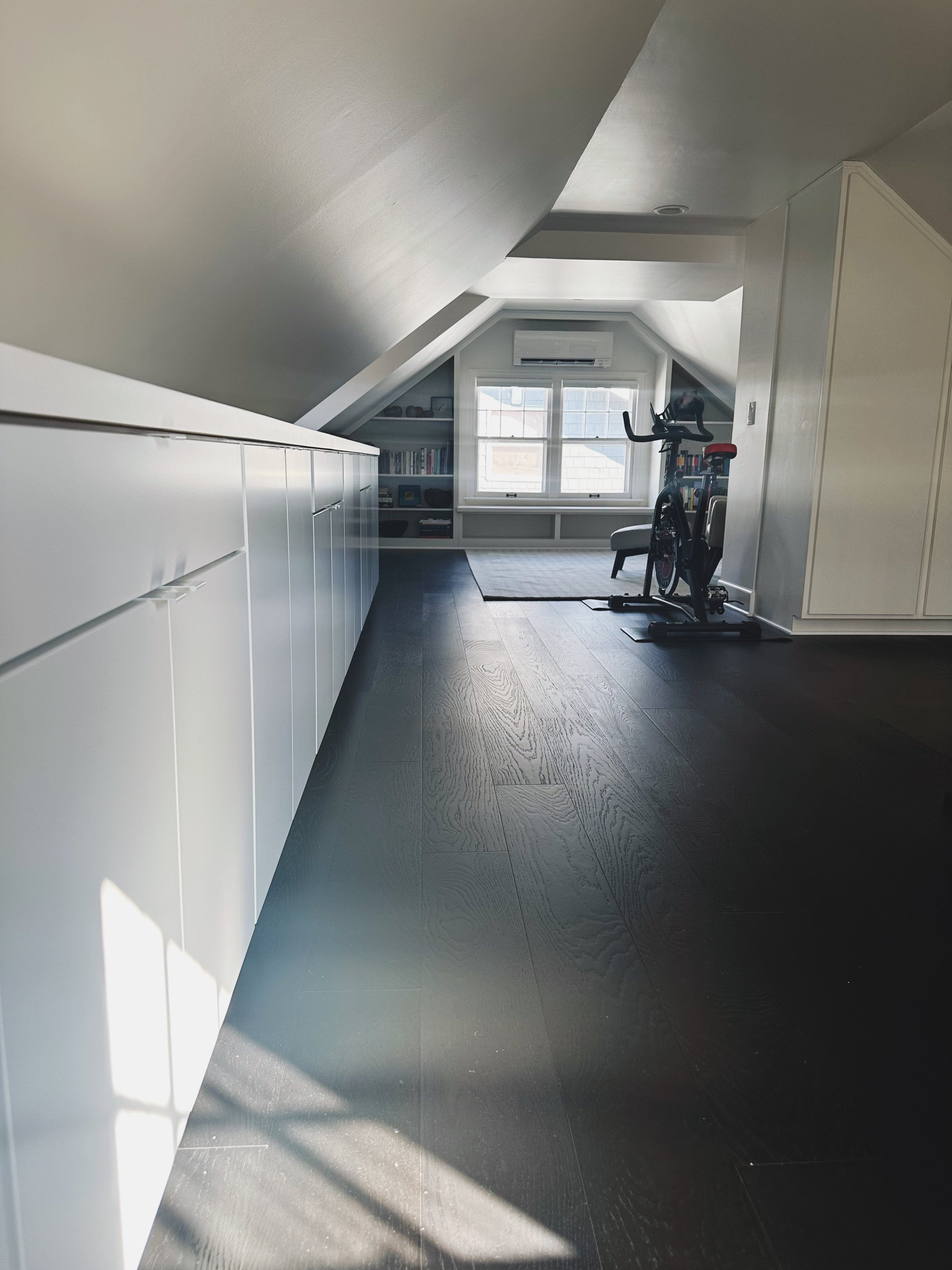A spacious attic room with a sloped ceiling, dark hardwood flooring, a large window, a desk with bookshelves, an exercise bike, and a chair.