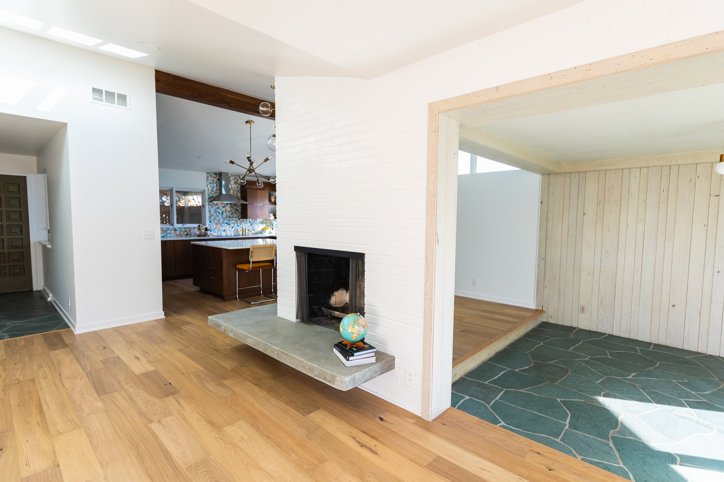 Living room with wooden flooring, white brick fireplace with a globe and books, open kitchen area with colorful backsplash, and a separate room with wood-paneled walls and slate flooring.