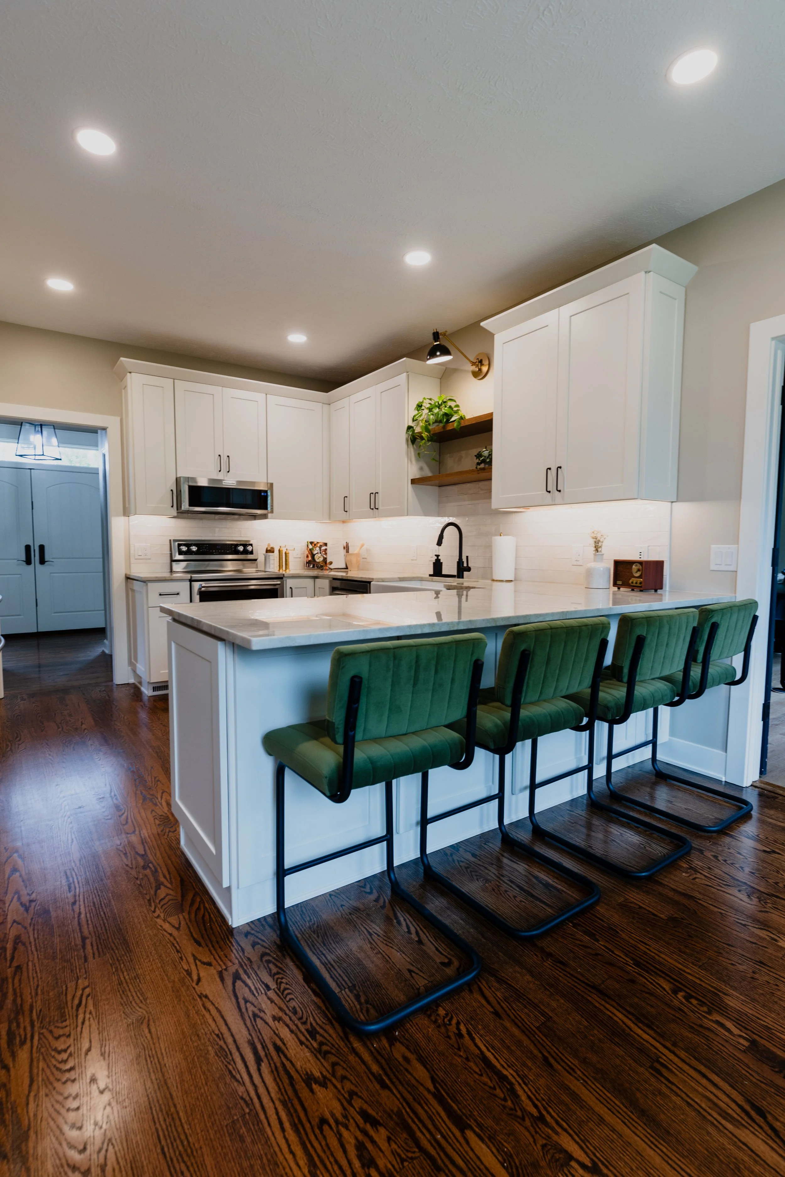 Modern kitchen with white cabinetry, marble countertop island, three green bar stools, hardwood flooring, and built-in appliances.